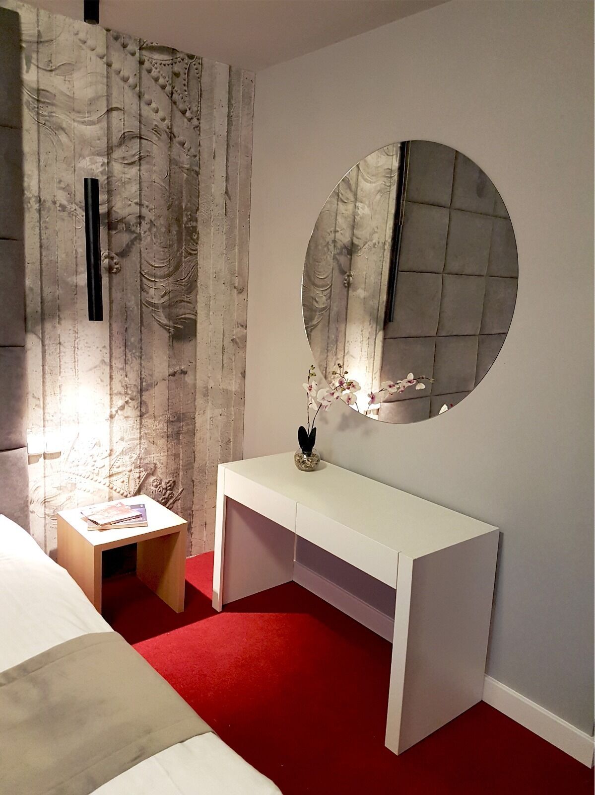 A bedroom with a bed, a white bedside table with a book, a white console bed, a round mirror, and a red carpet.