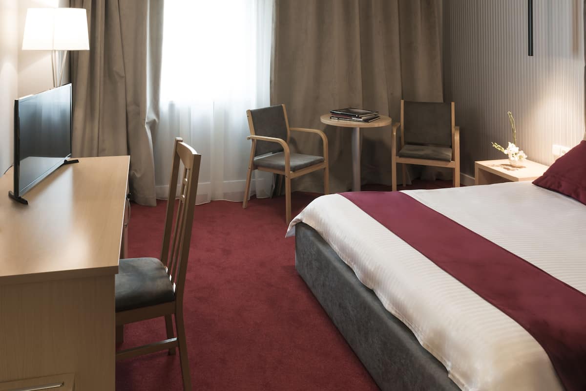 A hotel room with a bed, two chairs, a desk, a lamp, and a small table with a book and a vase of flowers.