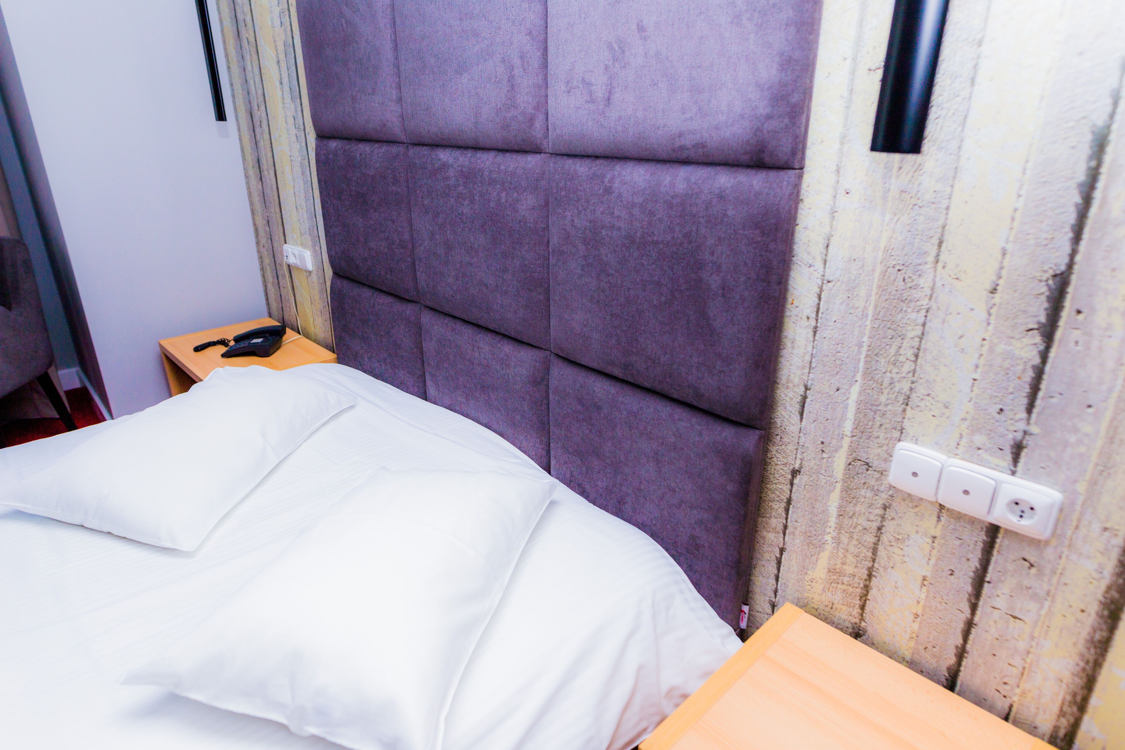A bed with a white comforter and pillows, a wooden nightstand with a black telephone, and a purple upholstered headboard.