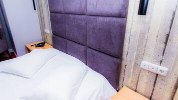 A bed with a white comforter and pillows, a wooden nightstand with a black telephone, and a purple upholstered headboard.
