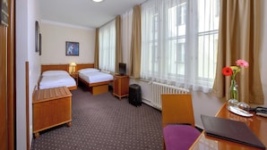 A hotel room with two beds, a desk with a laptop, a chair, a small table with a vase of flowers, a radiator, and a television on a nightstand. - Hotel Melantrich (Prague)