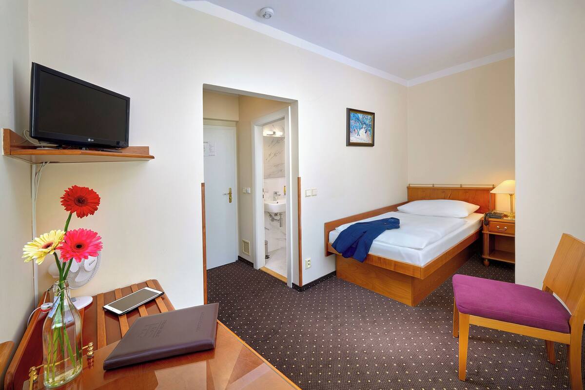 A hotel room with a bed, a desk with a laptop, a chair, a TV on a shelf, and a vase with flowers.