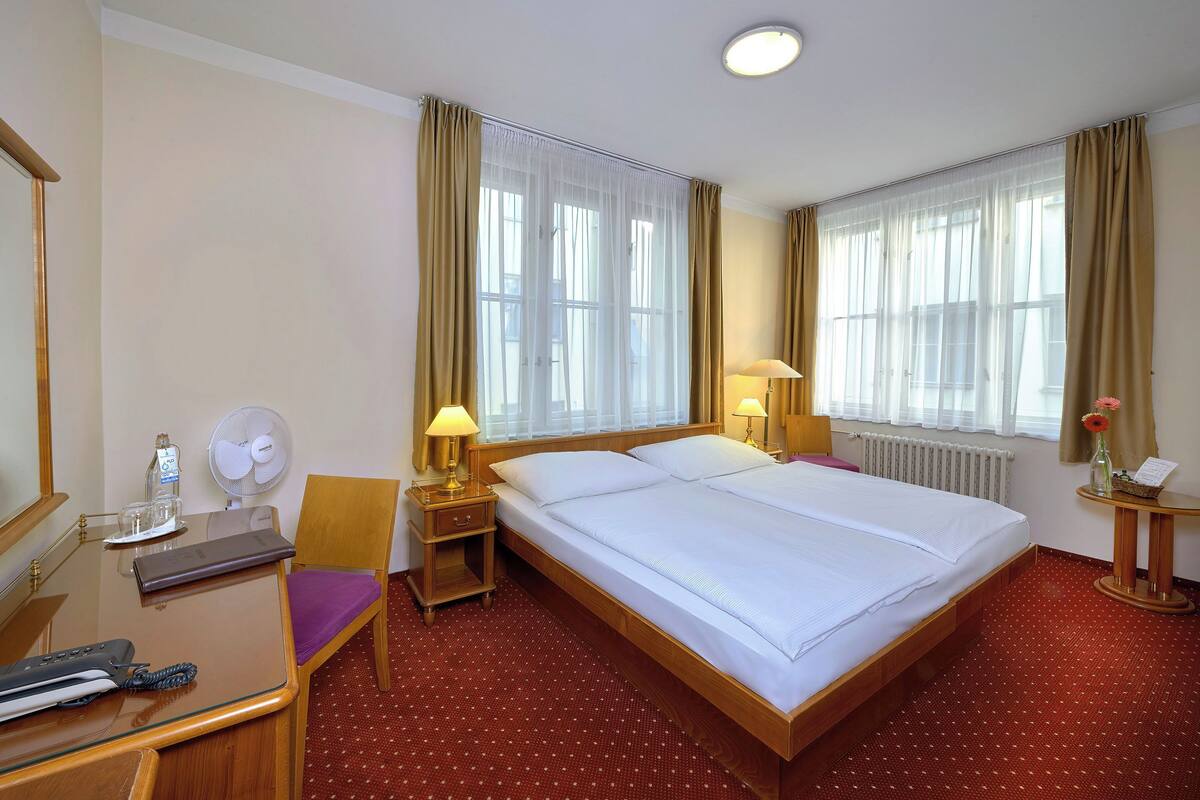 A hotel room with a large bed, a wooden nightstand with a lamp, a chair, a desk with a fan, and a small table with a vase.