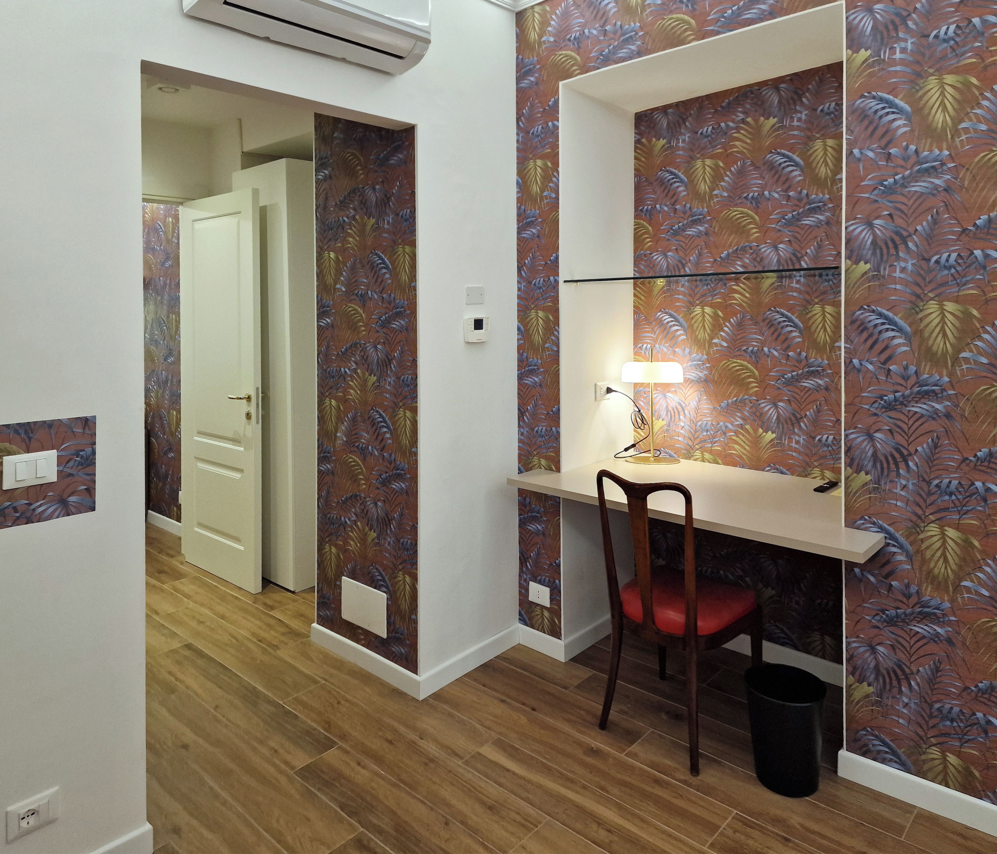 Ein Zimmer mit Schreibtisch, Stuhl und einer Wand mit Blätermuster. Eine Tür führt in einen weiteren Raum.