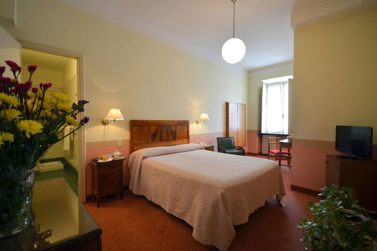 Una habitación de hotel con cama, mesitas de noche, escritorio con silla, televisor y ventana.