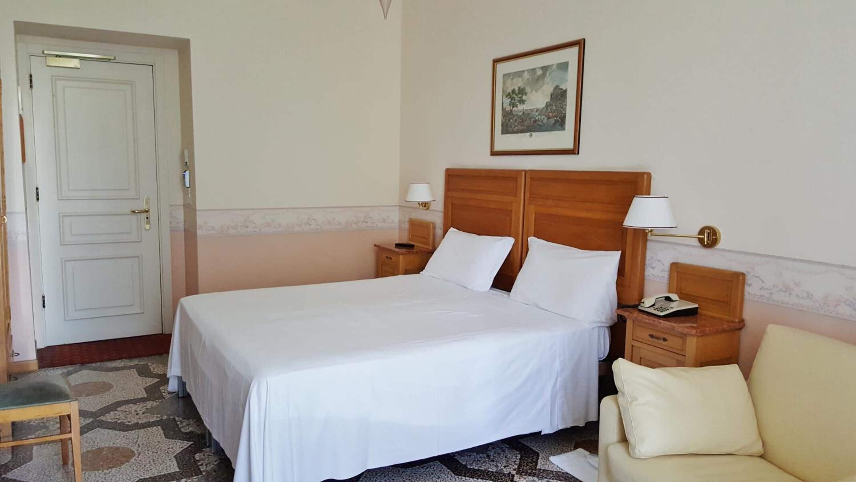 Une chambre d’hôtel avec un lit simple, deux tables de chevet avec des lampes, une tête de lit en bois, un tableau encadré au mur et un fauteuil beige.