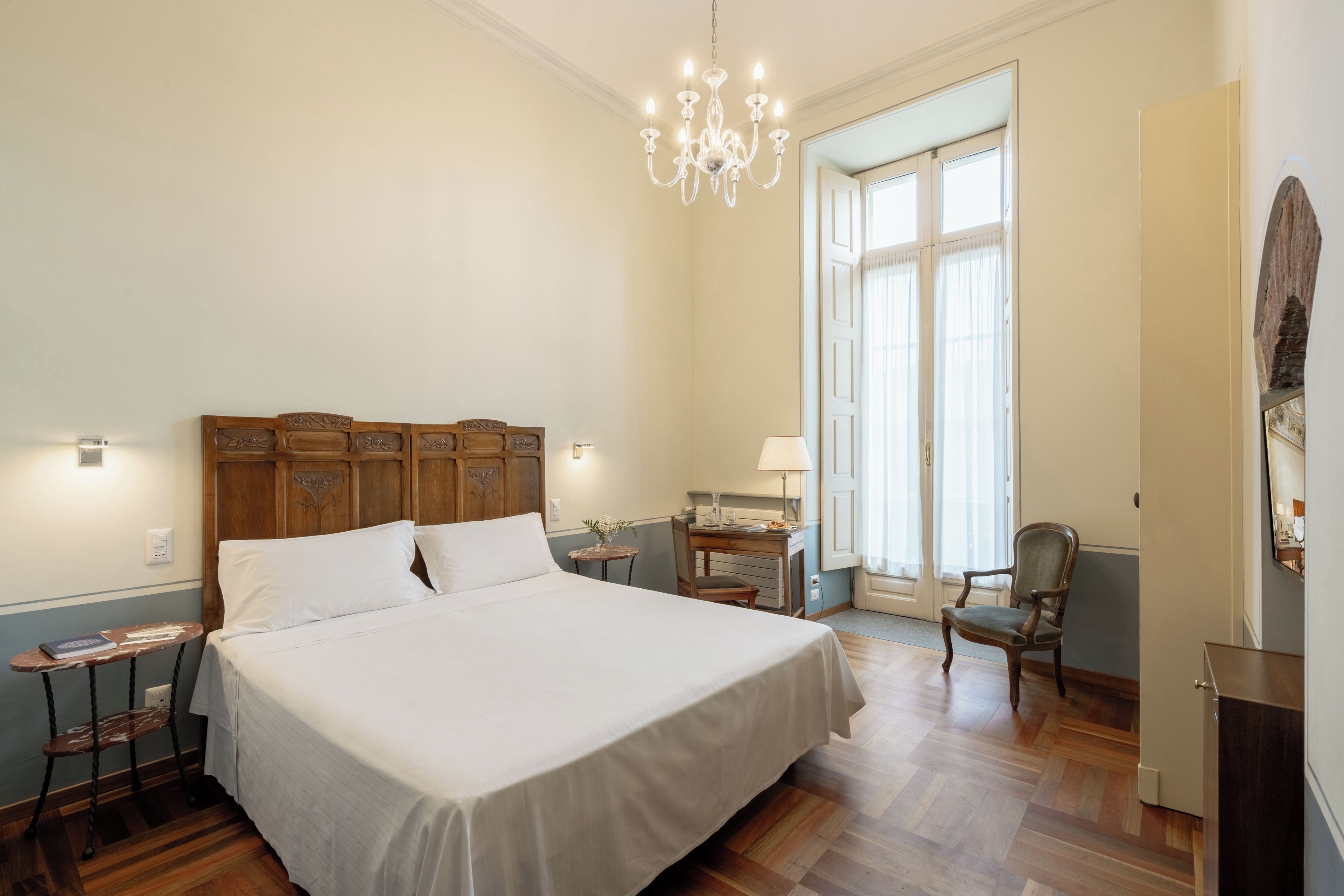 A bedroom with a large bed, a wooden headboard, a chandelier, a desk with a chair, and a window with curtains.