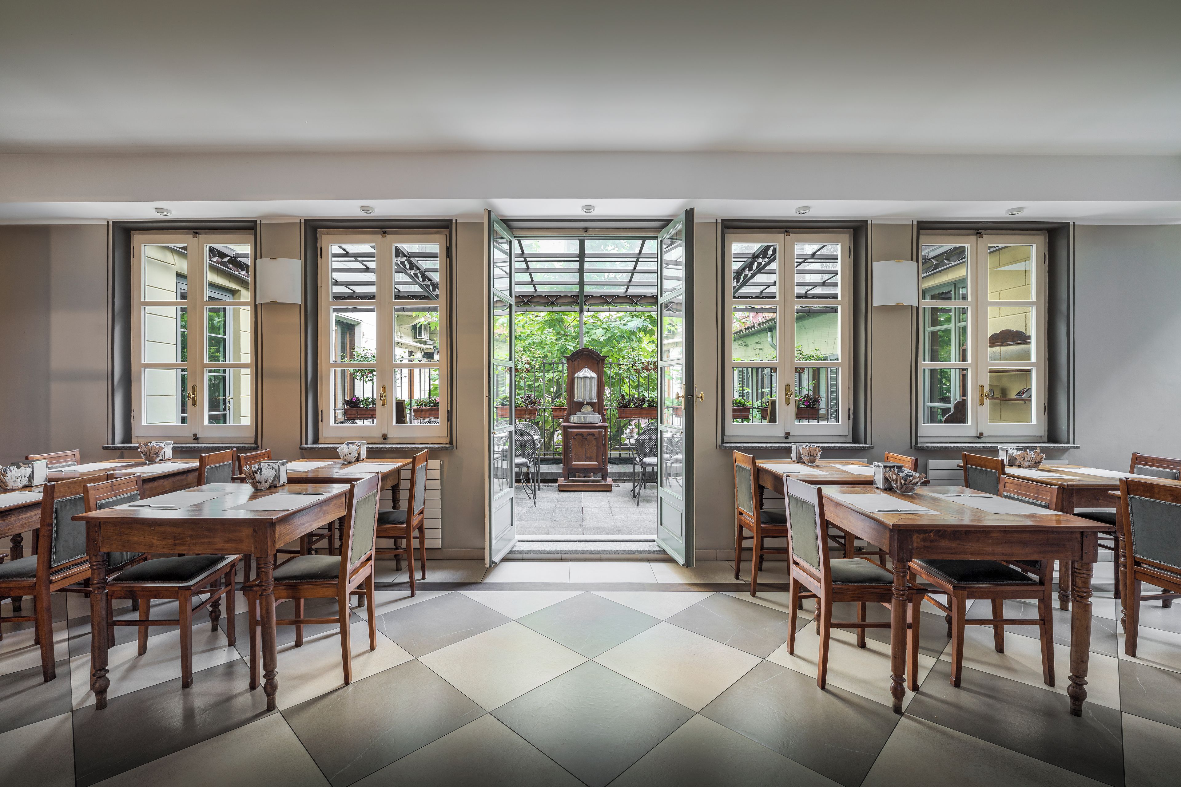 Un espace repas comprenant plusieurs tables et chaises, de grandes fenêtres donnant sur un paysage verdoyant et une porte centrale ouverte qui mène à l’extérieur.