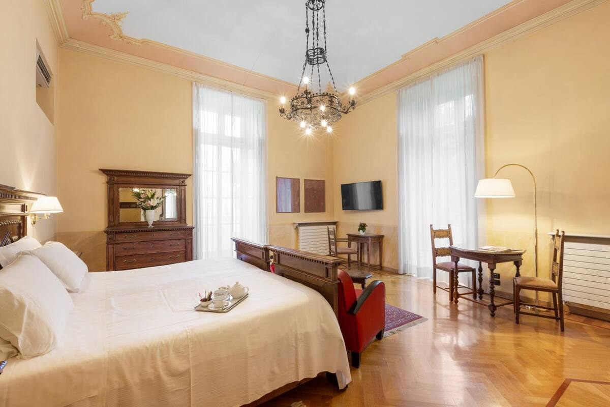 Habitación de hotel con una cama grande, un aparador de madera, un escritorio con dos sillas, un televisor y una araña.