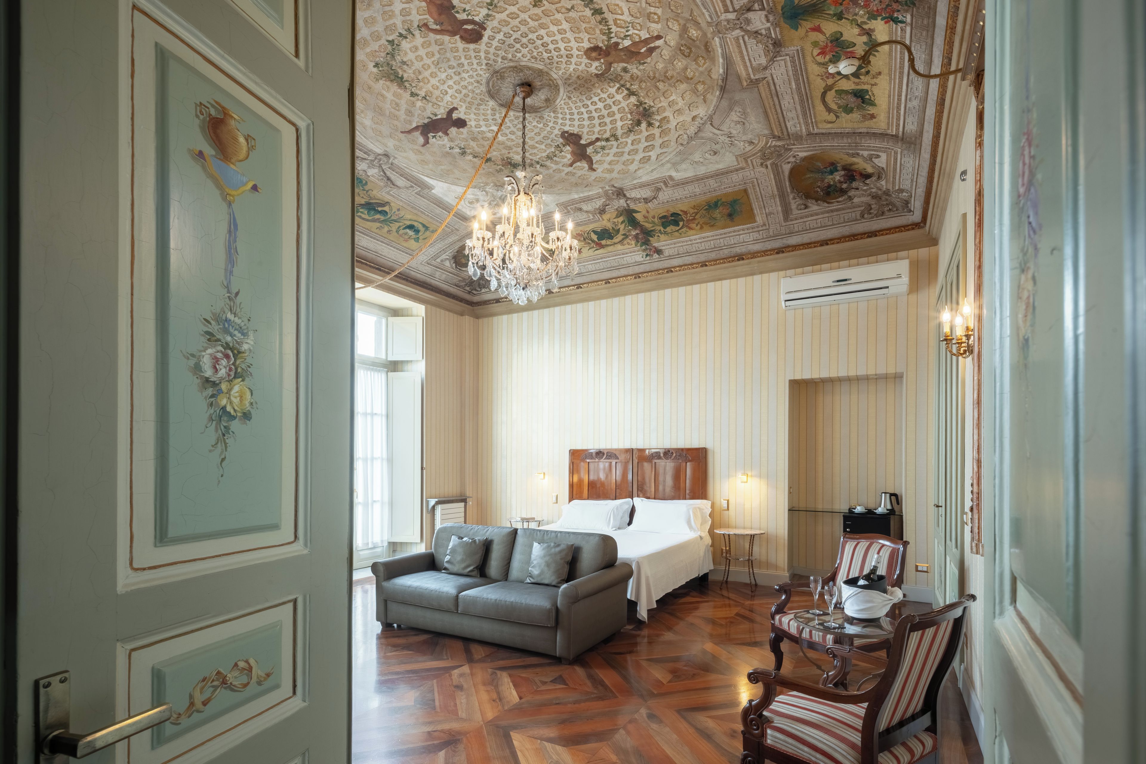 A hotel room with a large bed, a sofa, a chandelier, and a decorative ceiling.