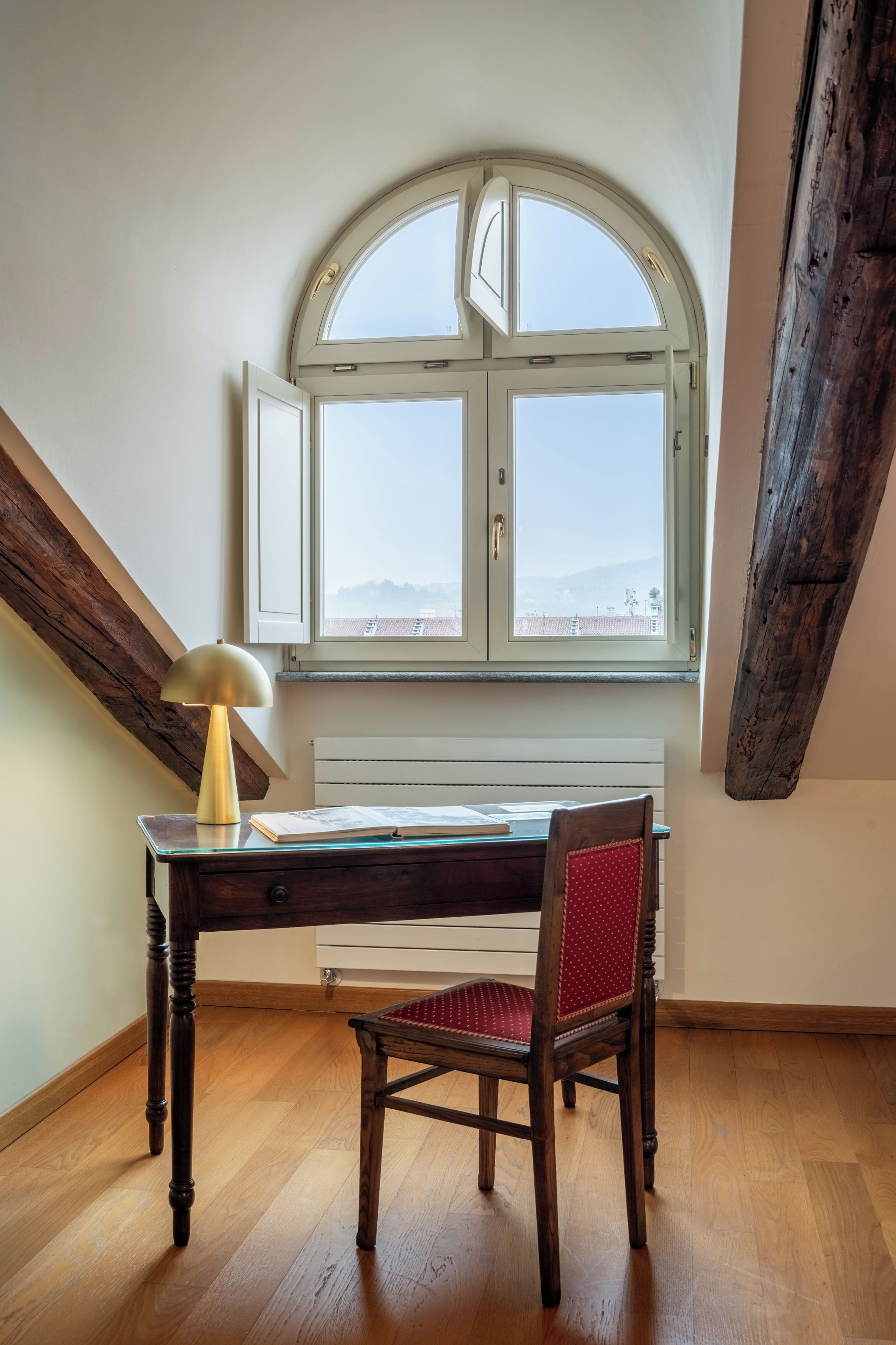 A room with a wooden desk, a chair, a lamp, and a large arched window.