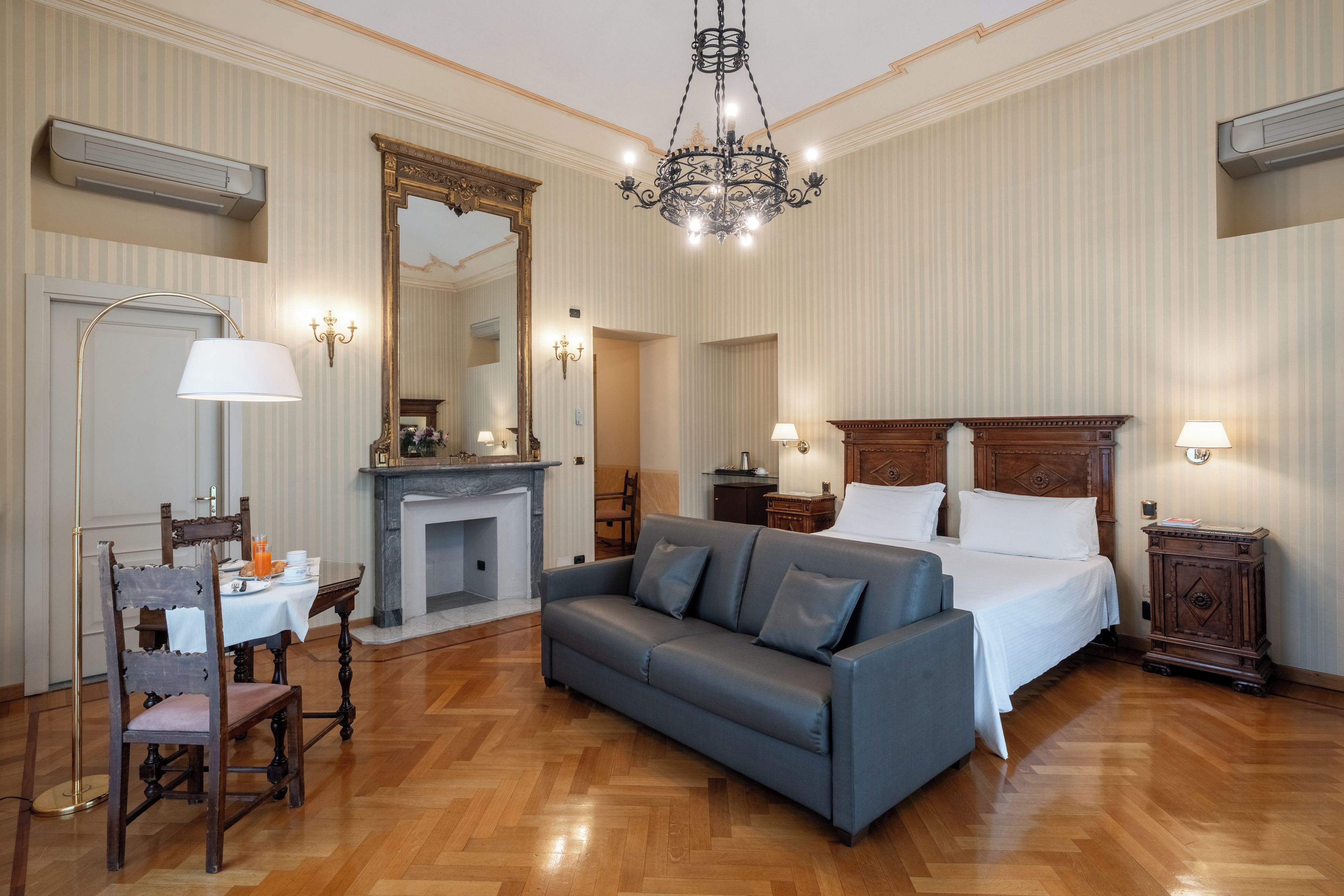 A hotel room with a large bed, a sofa, a dining area with a table and chairs, a fireplace, and a chandelier.