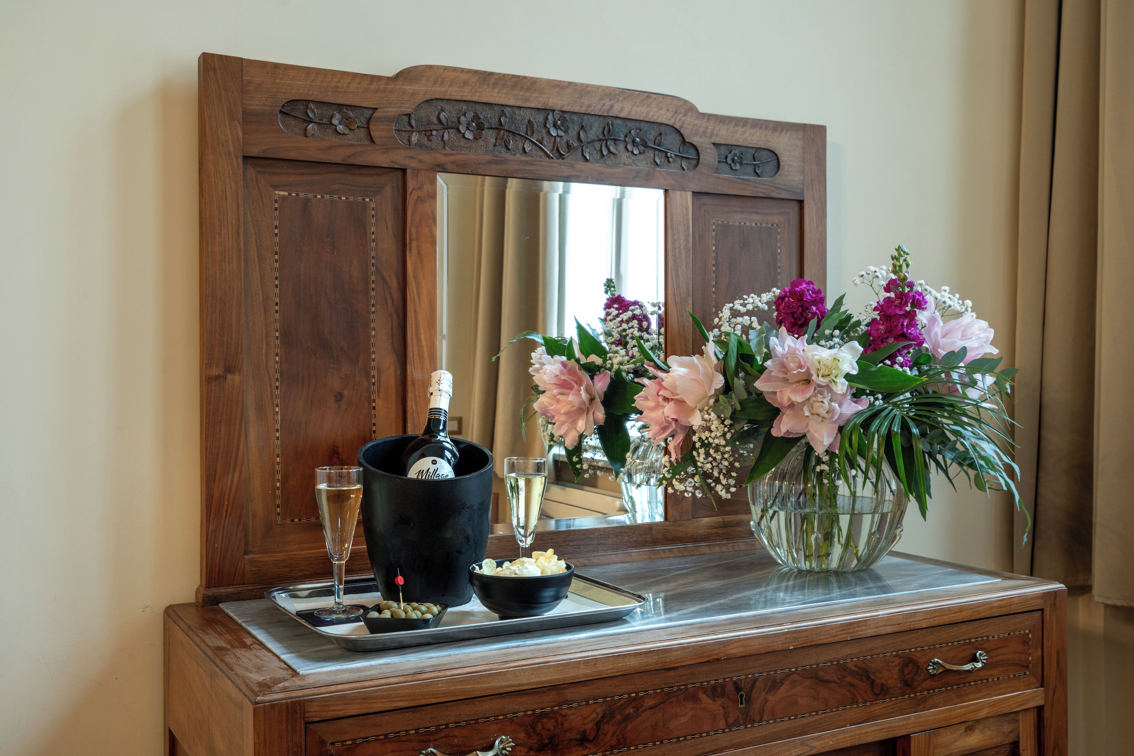Een houten dressoir met een spiegel, een champagnefles, glazen en een bloemstuk.