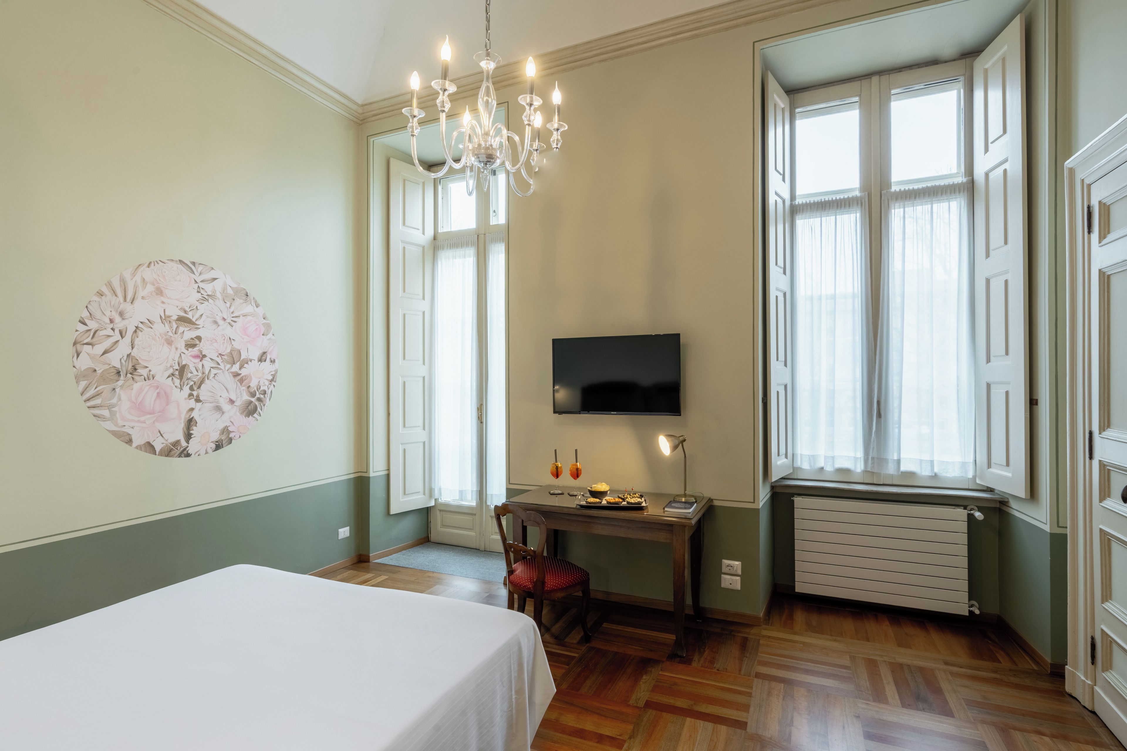 A hotel room with a bed, a desk with a computer, a TV, a radiator, and a chandelier.