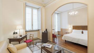 Una camera d'albergo con un letto, un'area salotto con tavolo e sedia, una piccola scrivania con lampada e una finestra con tende.