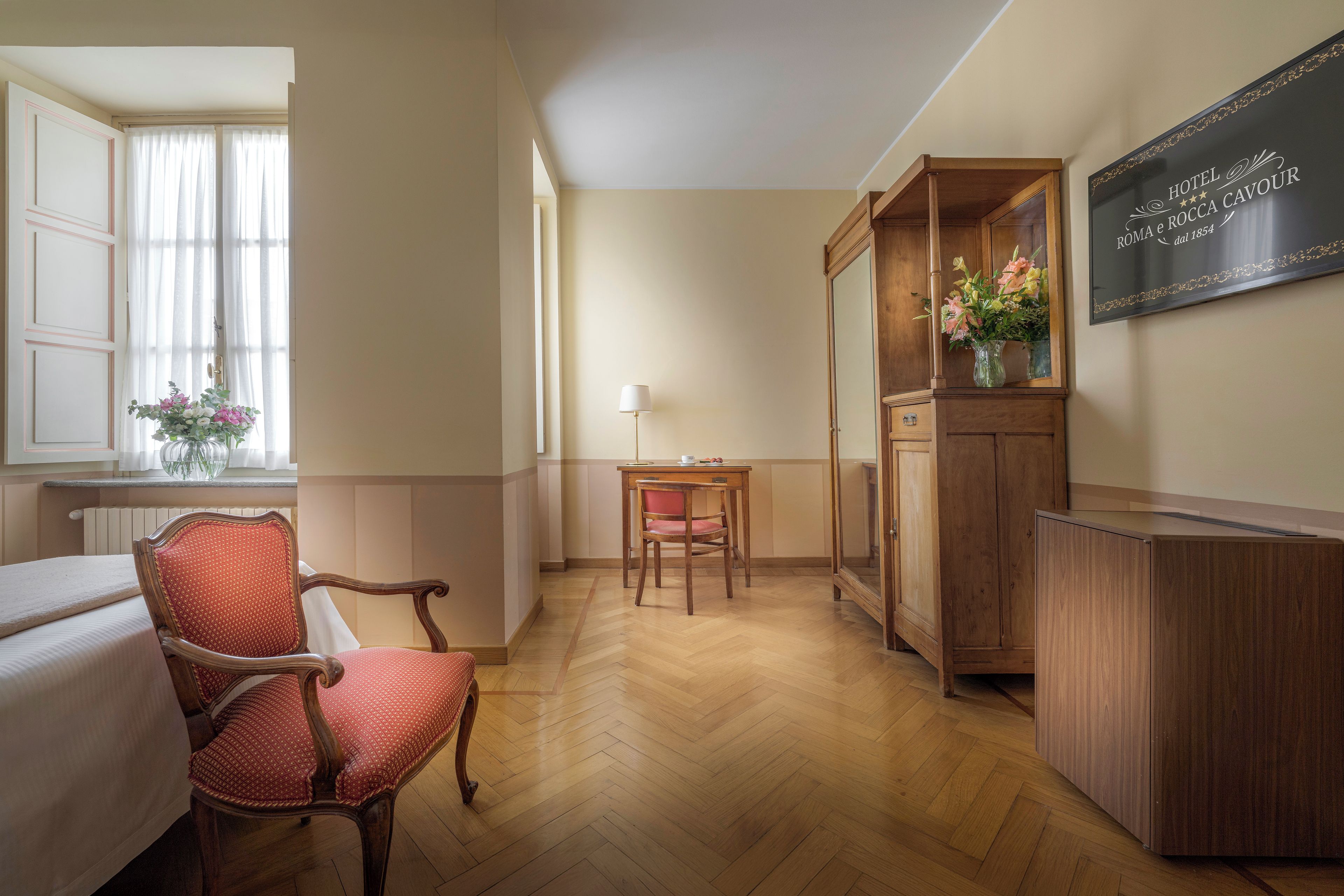 A hotel room with a wooden floor, a dining table with chairs, a cabinet with a mirror, and a window with curtains.