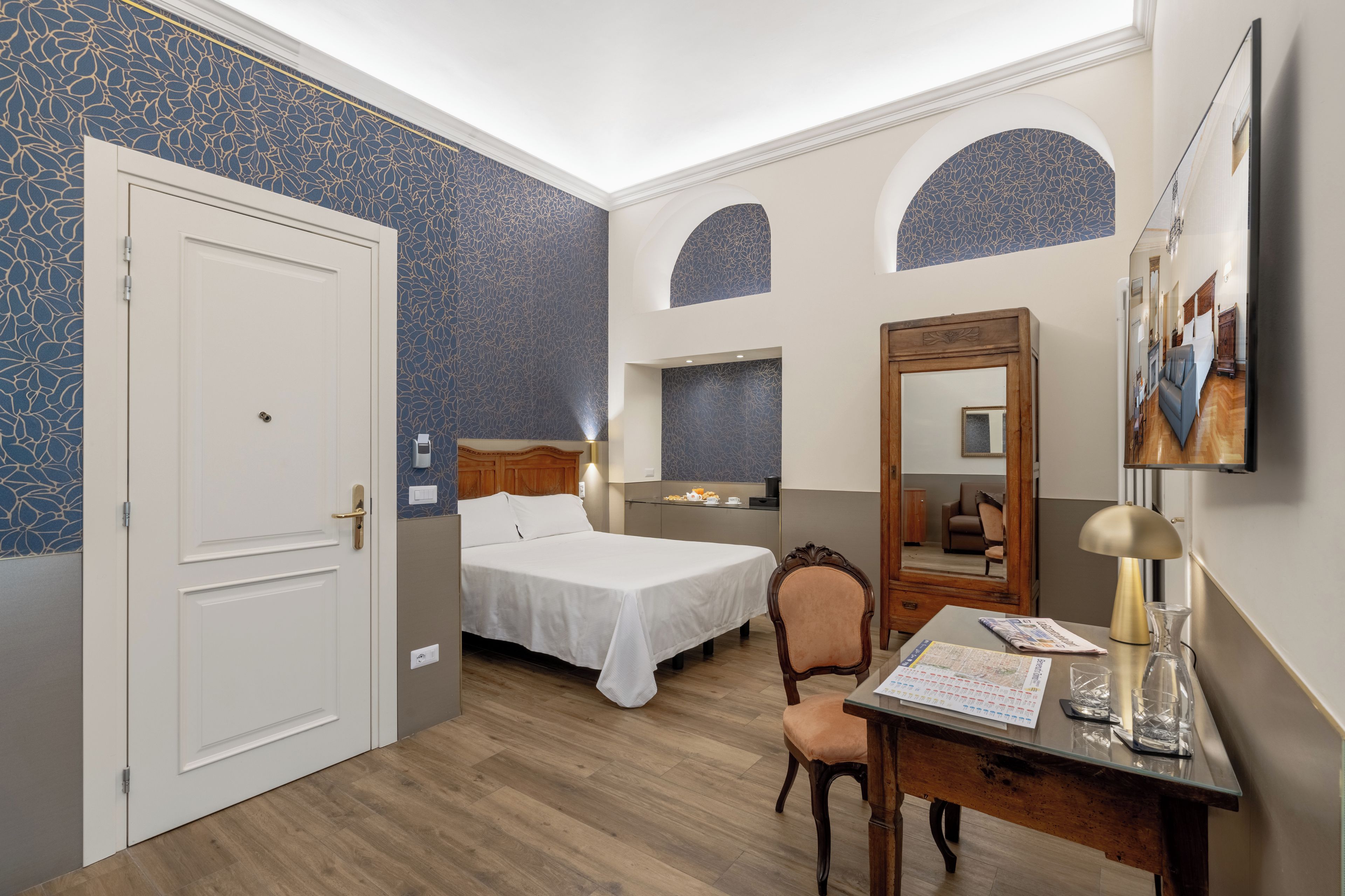 A hotel room with a bed, a desk, a chair, a mirror, and a wall with a blue floral pattern.