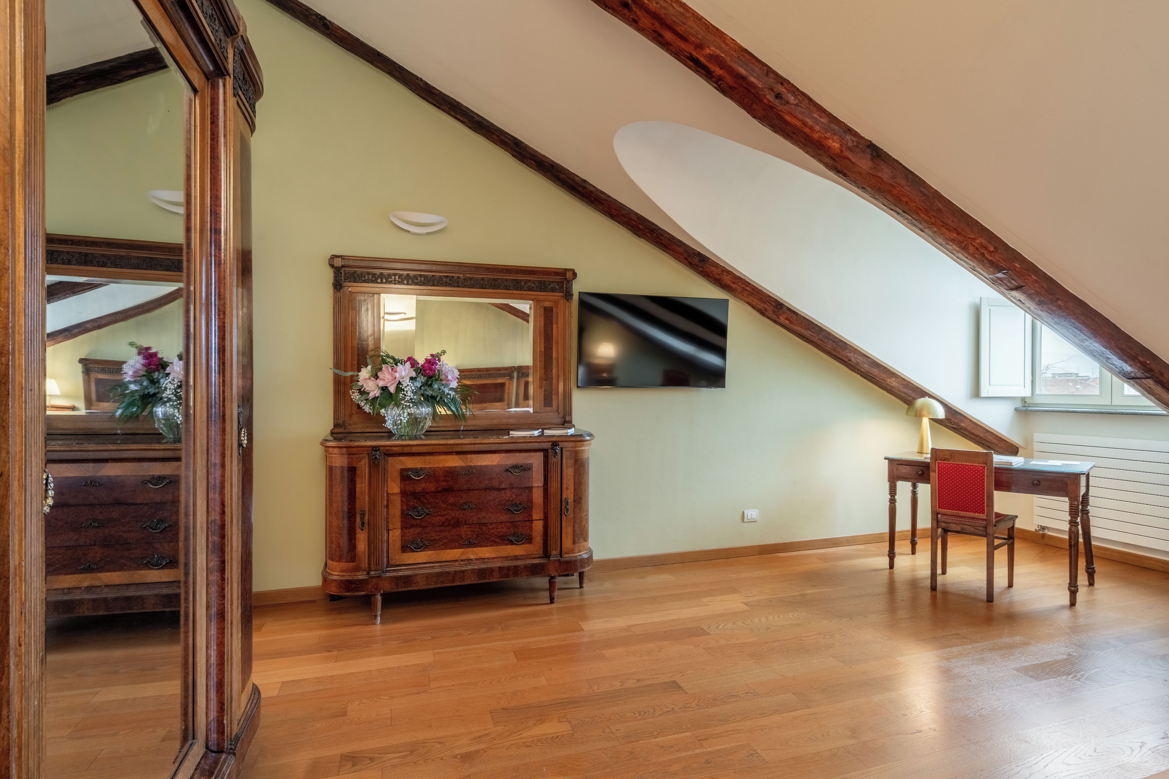 Una habitación con piso de madera, un espejo grande, un mueble de madera con cajones, un florero con flores, un escritorio con una silla y un televisor montado en la pared.