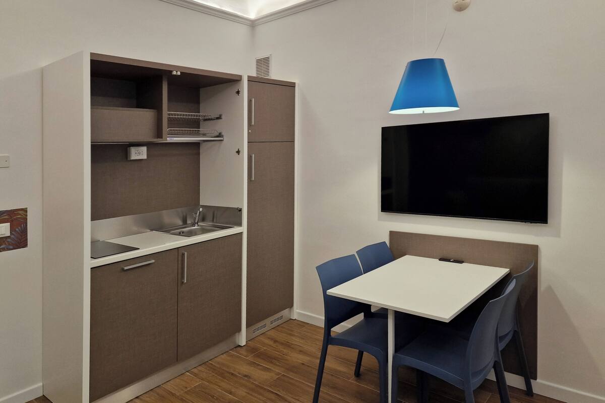 Una cocina compacta con estufa integrada, lavaplatos y armarios de almacenamiento. Un comedor con sillas está ubicado debajo de un televisor grande montado en la pared.