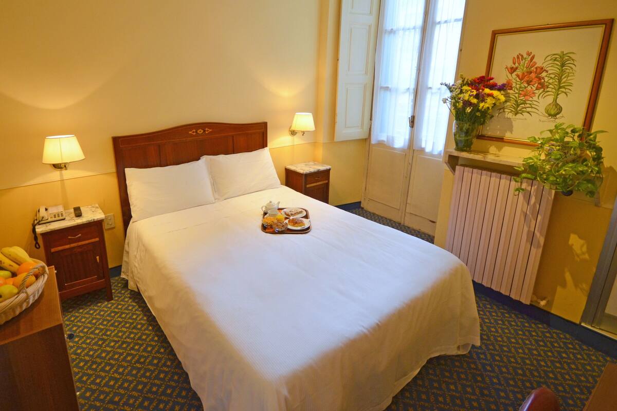 Habitación de hotel con una cama bien hecha, una cesta de frutas, una mesita de noche con lámpara y una ventana con vistas al exterior.
