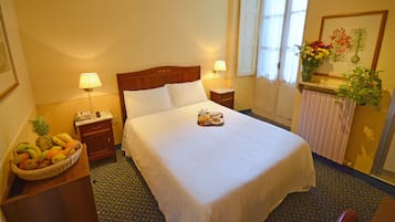 Ein Hotelzimmer mit einem ordentlich bezogenen Bett, einem Obstkorb, einem Nachttisch mit Lampe und einem Fenster mit Blick ins Freie.