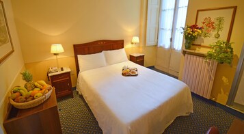 Ein Hotelzimmer mit einem ordentlich bezogenen Bett, einem Obstkorb, einem Nachttisch mit Lampe und einem Fenster mit Blick ins Freie.