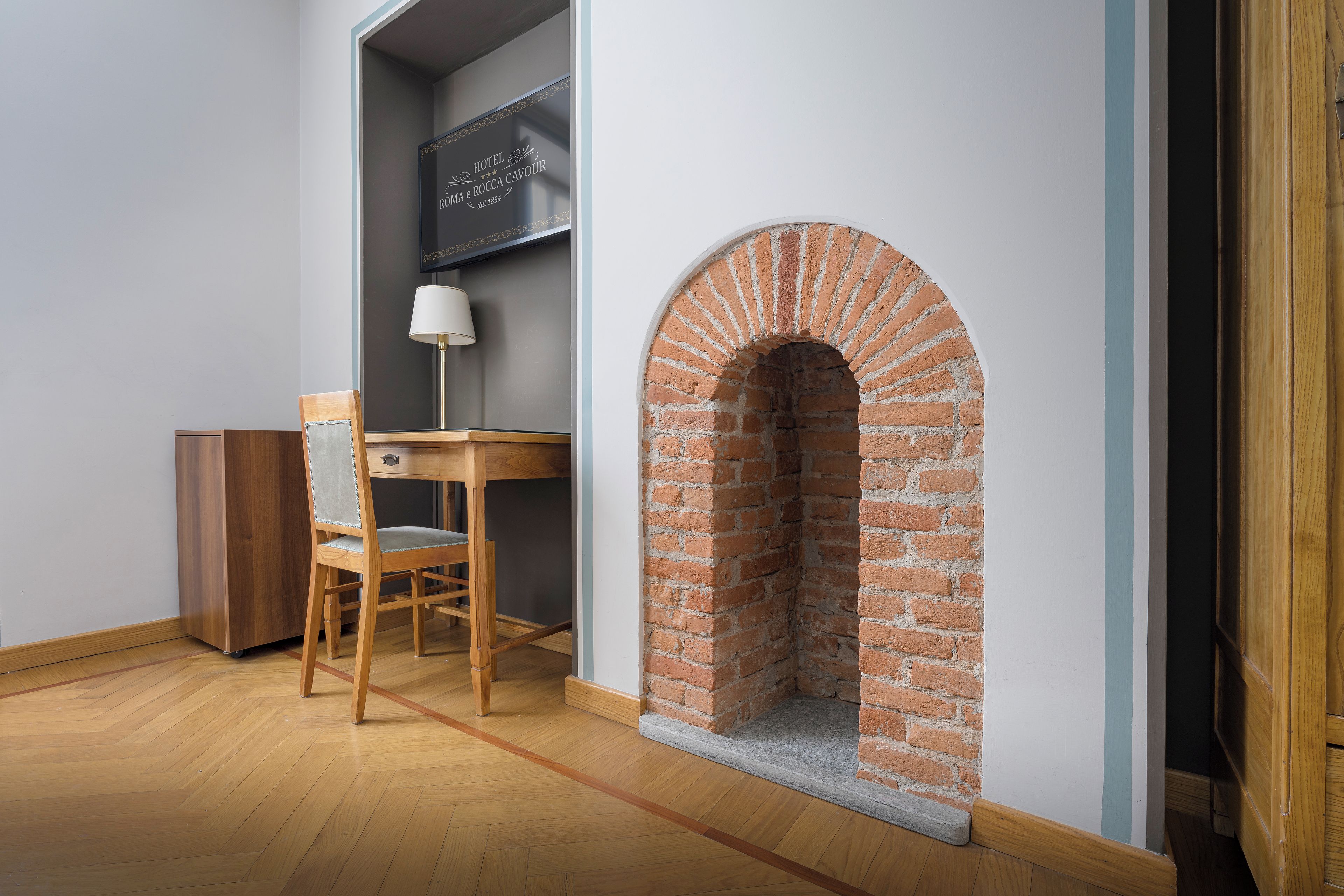 A room with a brick fireplace, a wooden desk with a chair, and a lamp.