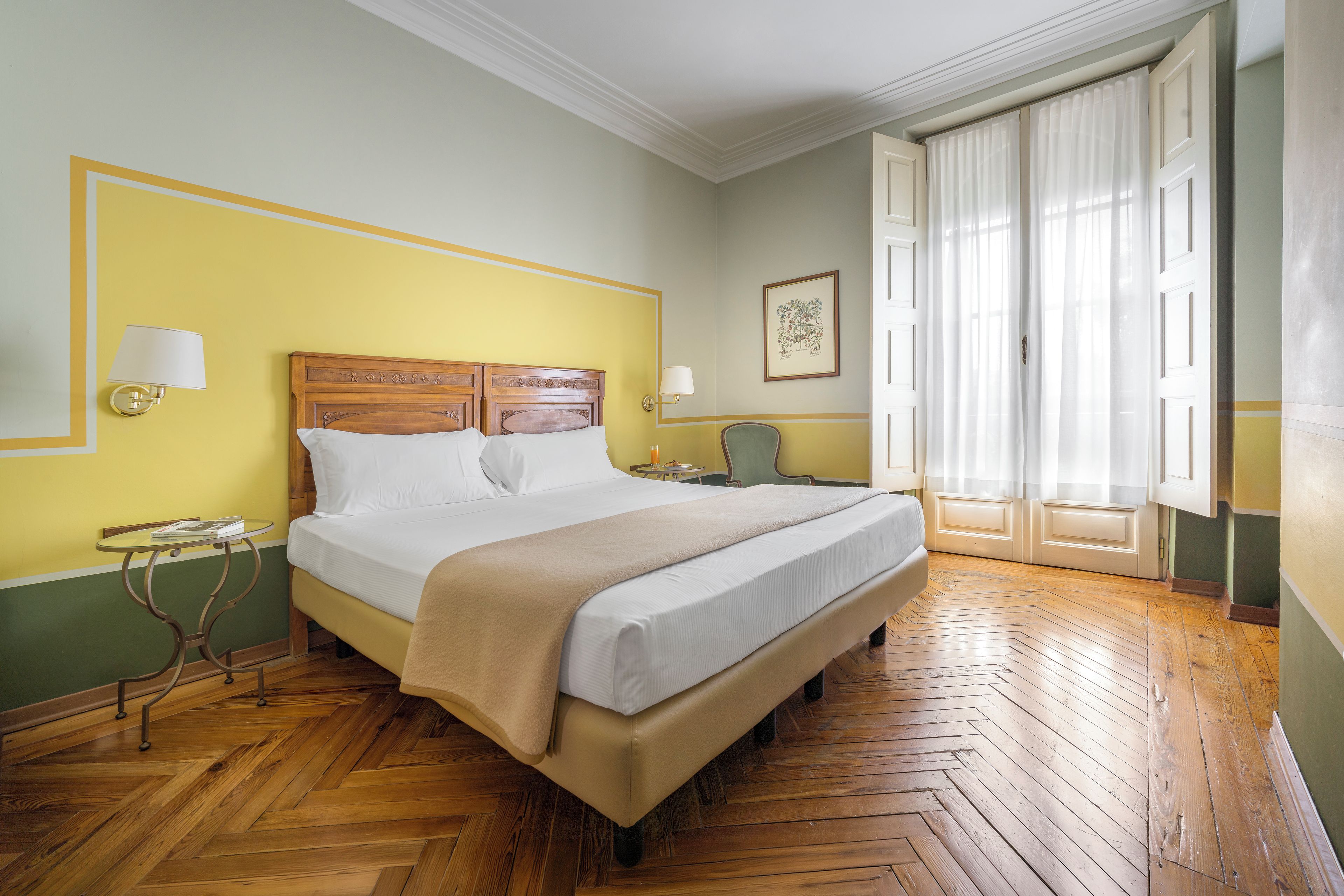 Una habitación de hotel con una cama grande, una mesita de noche, una silla y un ventanal con cortinas.