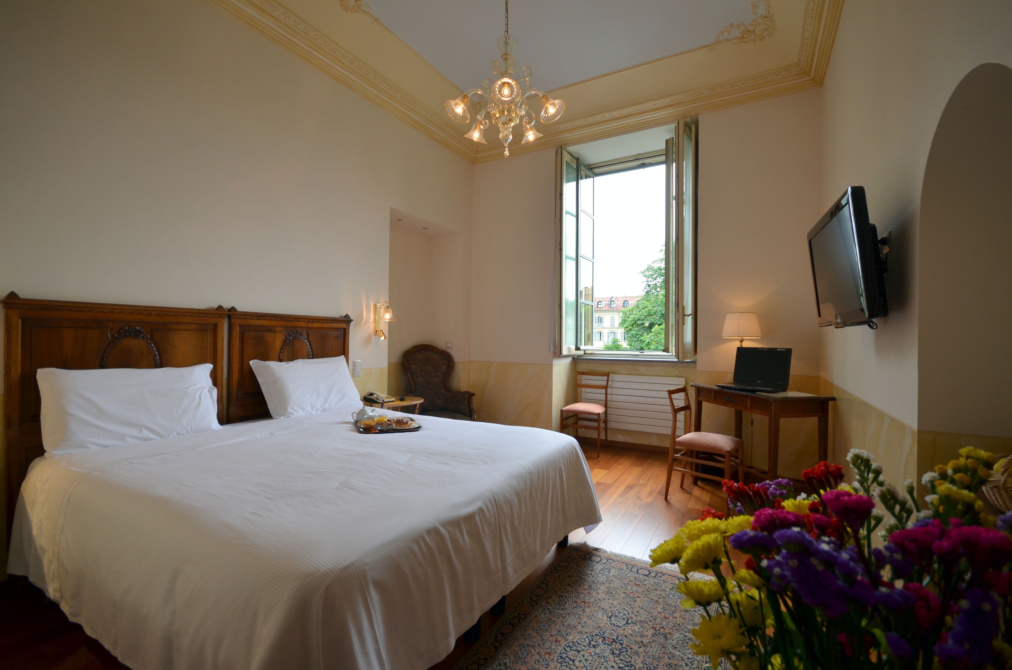 Un dormitorio con una cama grande, un escritorio con computadora, televisor en la pared y ventana con vista a edificios y árboles.