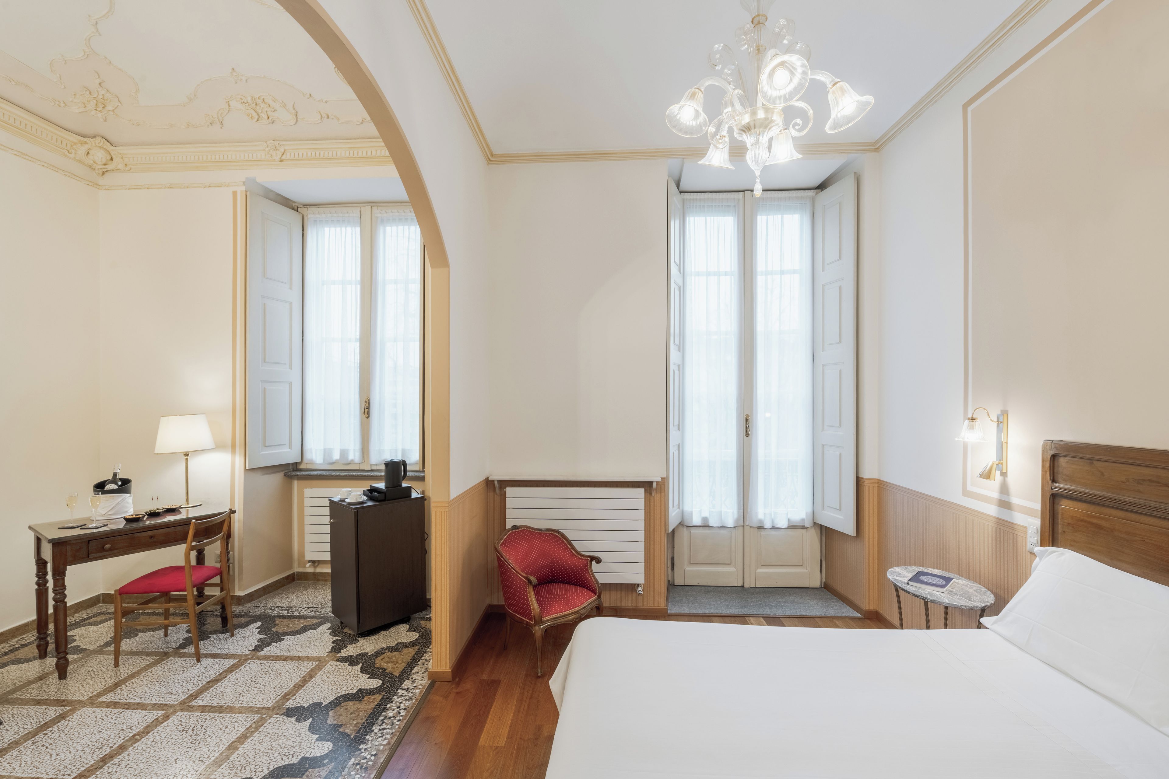 Ein Hotelzimmer mit einem Bett, einem Schreibtisch mit Stuhl, einem roten Sessel, einem kleinen Tisch mit Lampe und einer Kandelaber.