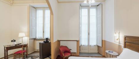 Ein Hotelzimmer mit einem Bett, einem Schreibtisch mit Stuhl, einem roten Sessel, einem kleinen Tisch mit Lampe und einer Kandelaber.
