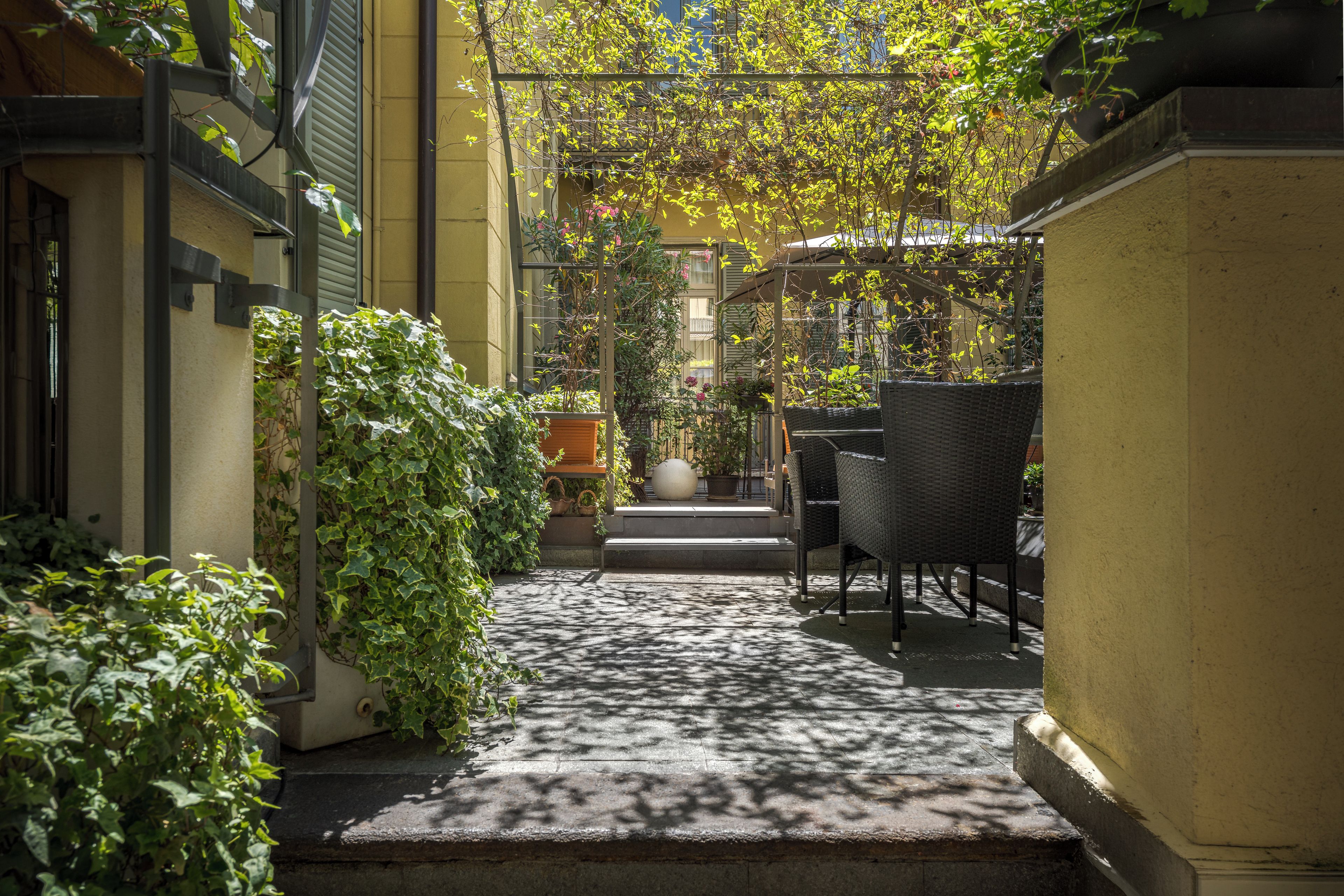 Un étroit couloir extérieur doté d’un sol en béton, bordé de verdure et agrémenté d’une chaise en osier.