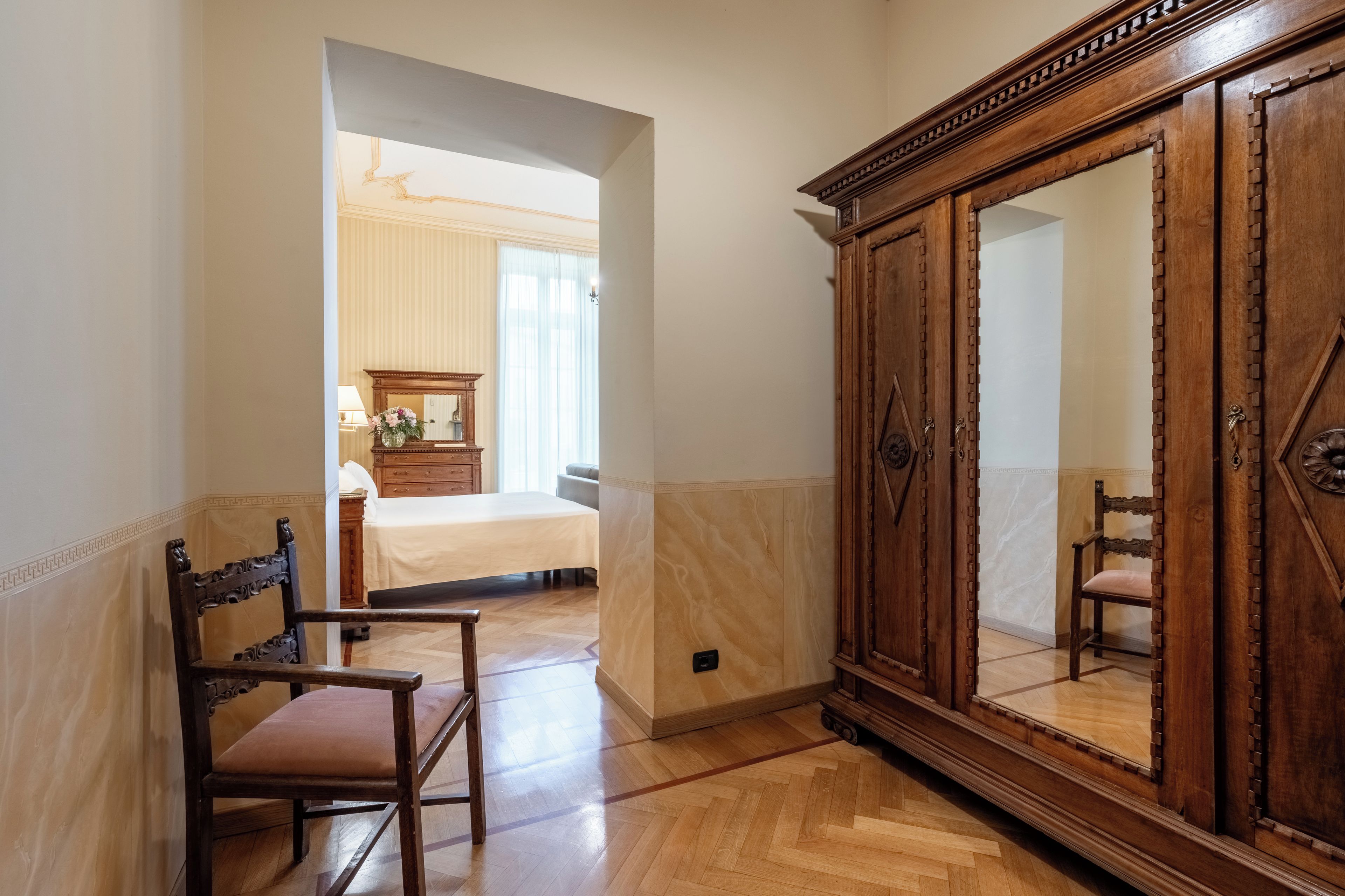 A hotel room with a wooden wardrobe, a bed, a chair, and a mirror.