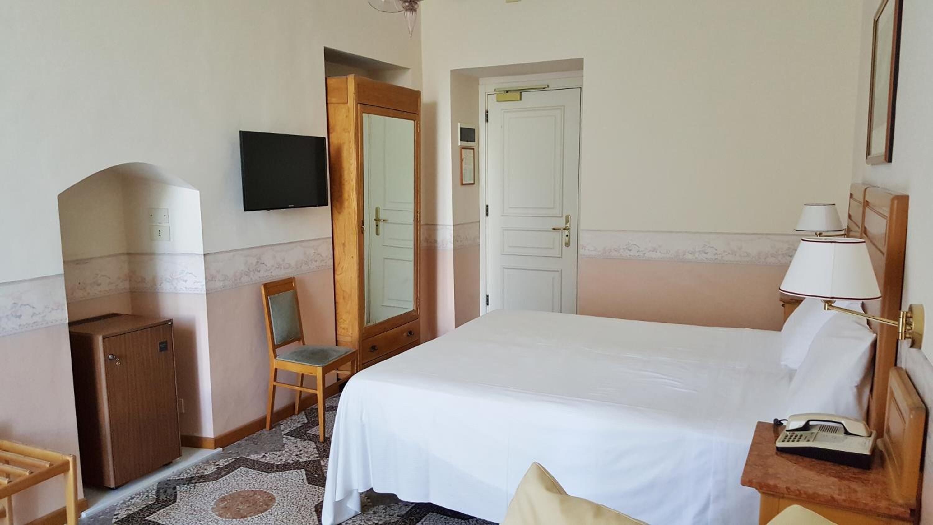 Habitación de hotel con cama, escritorio con lámpara, silla, televisor y armario.