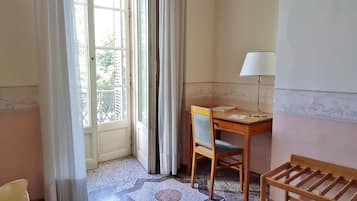 Habitación con escritorio, silla y lámpara. Cuenta con una ventana con cortinas y vistas al exterior con vegetación.