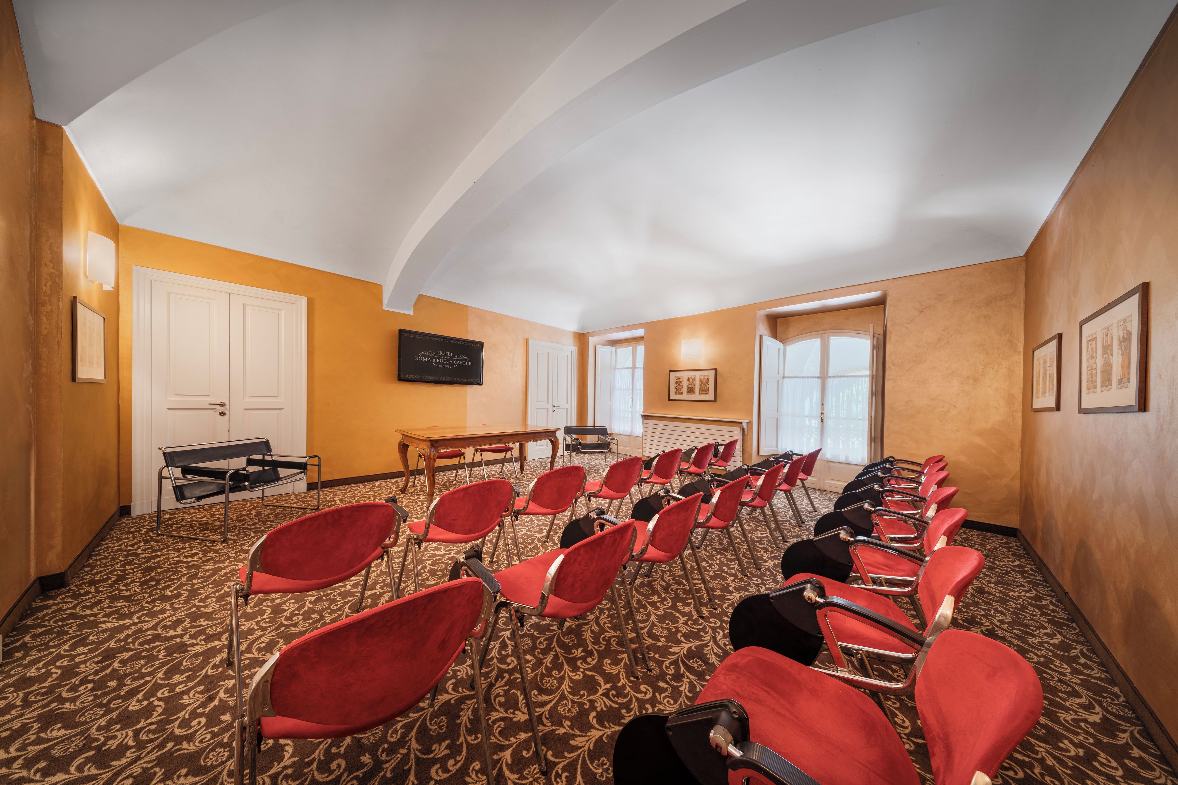 Un salón de conferencias con filas de sillas rojas, una mesa de madera y una puerta blanca.