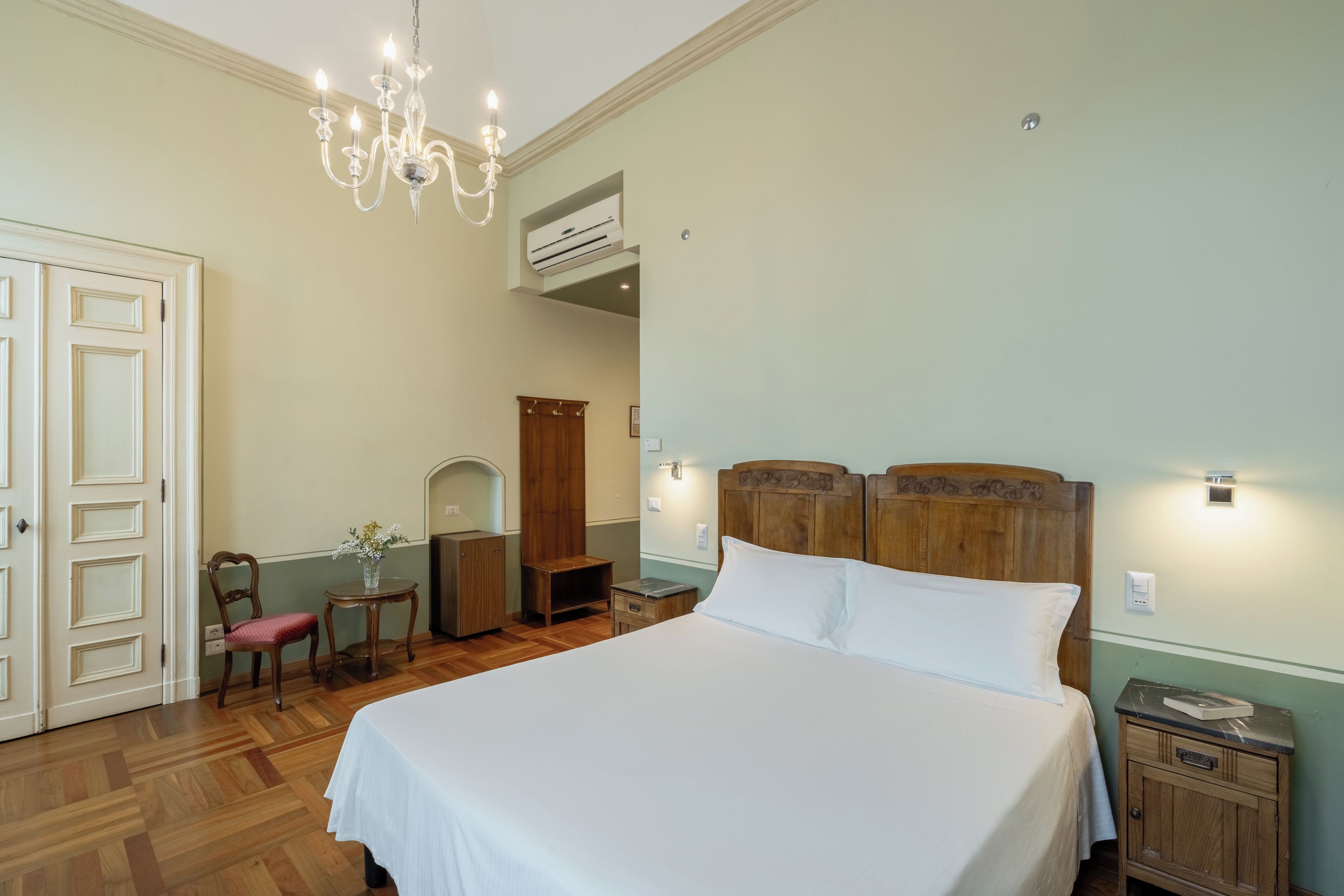 Un dormitorio con una cama grande, una mesita de noche de madera, una silla, una pequeña mesa con un florero y una araña.