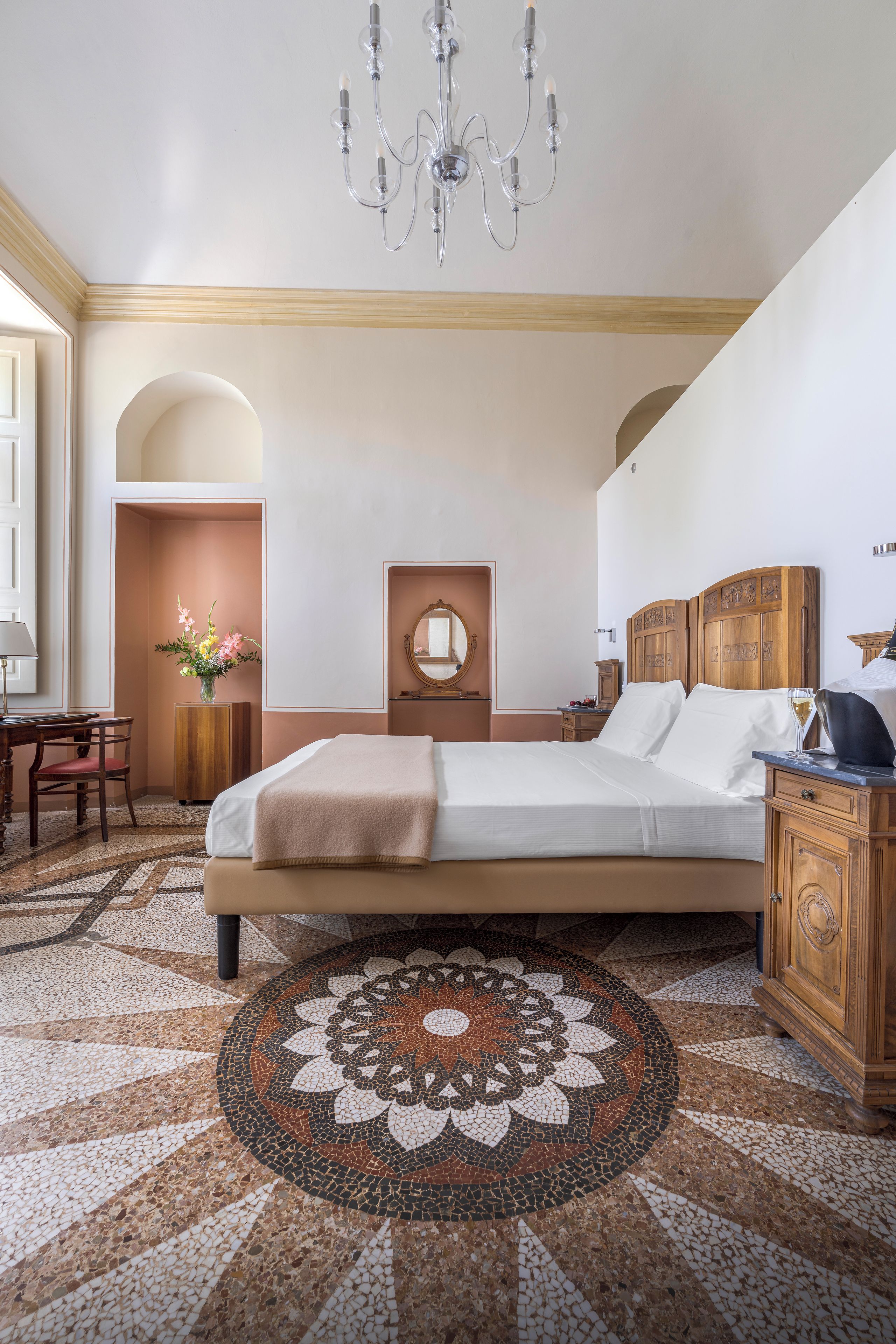 A hotel room with a large bed, a wooden headboard, a round mirror, a desk with a chair, and a chandelier.