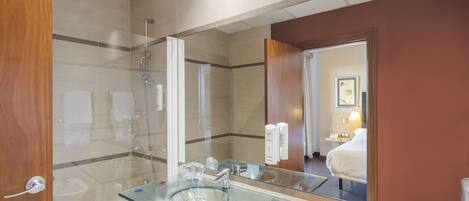 A modern bathroom with a glass sink, a shower, and a large mirror reflecting a bedroom with a bed and a nightstand.