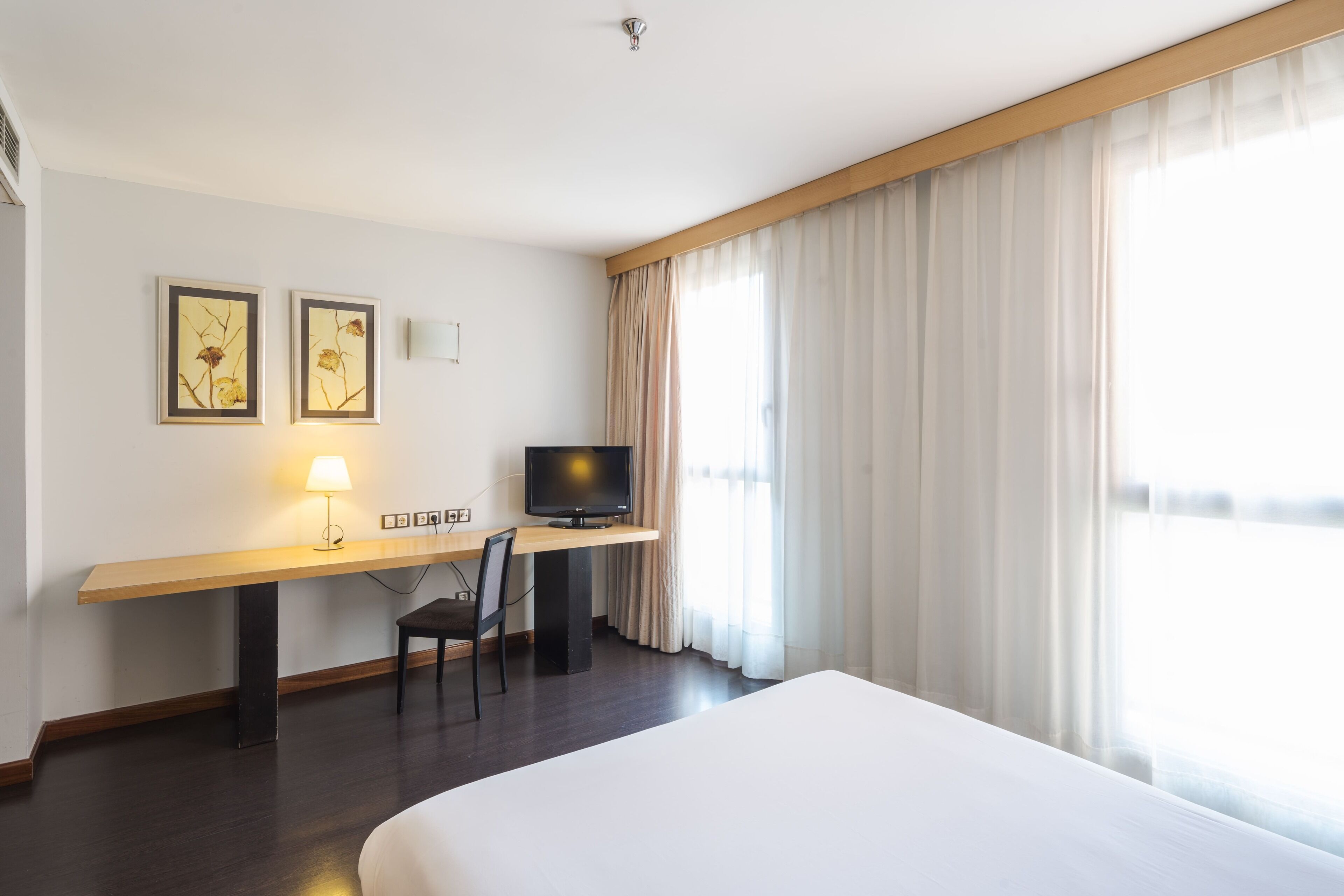 A hotel room with a large bed, a desk with a television, two framed pictures, and a lamp.