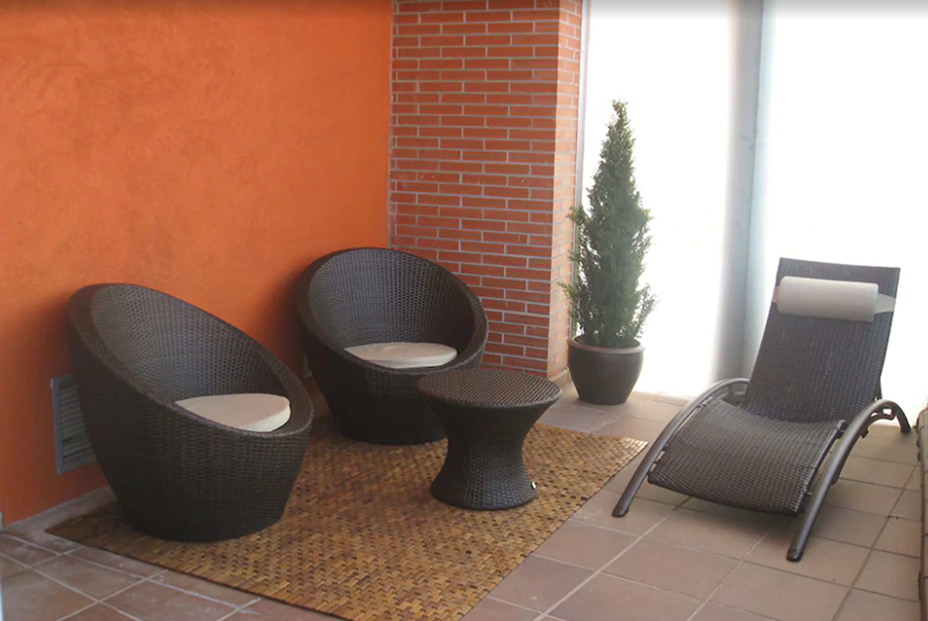 A balcony with wicker furniture, including two chairs and a small table, a potted plant, and a view of the outside.
