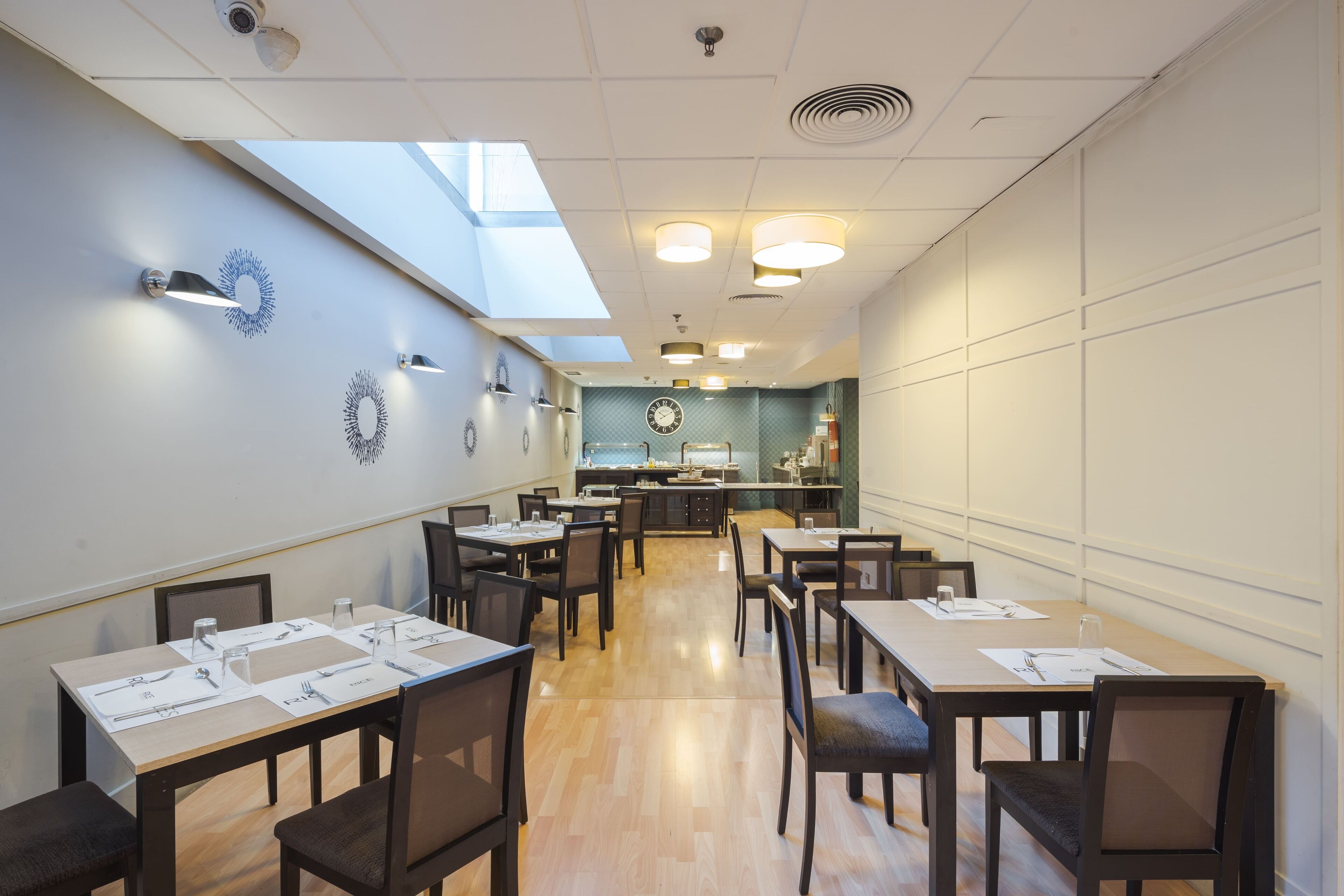 A restaurant with a high ceiling, skylight, and a kitchen in the background. Tables are set with menus and cutlery.