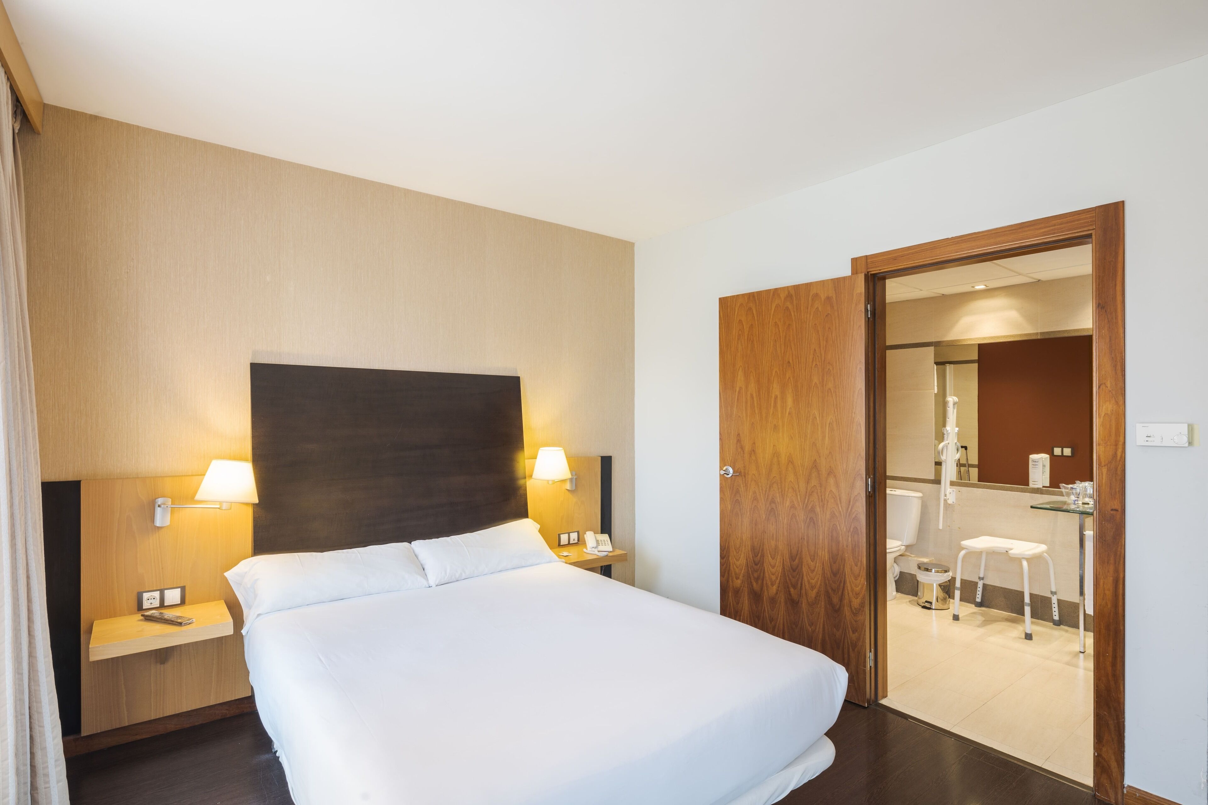 Habitación de hotel con cama, lámparas de noche, cabecera de madera, puerta de madera y un baño visible a través de una puerta abierta.
