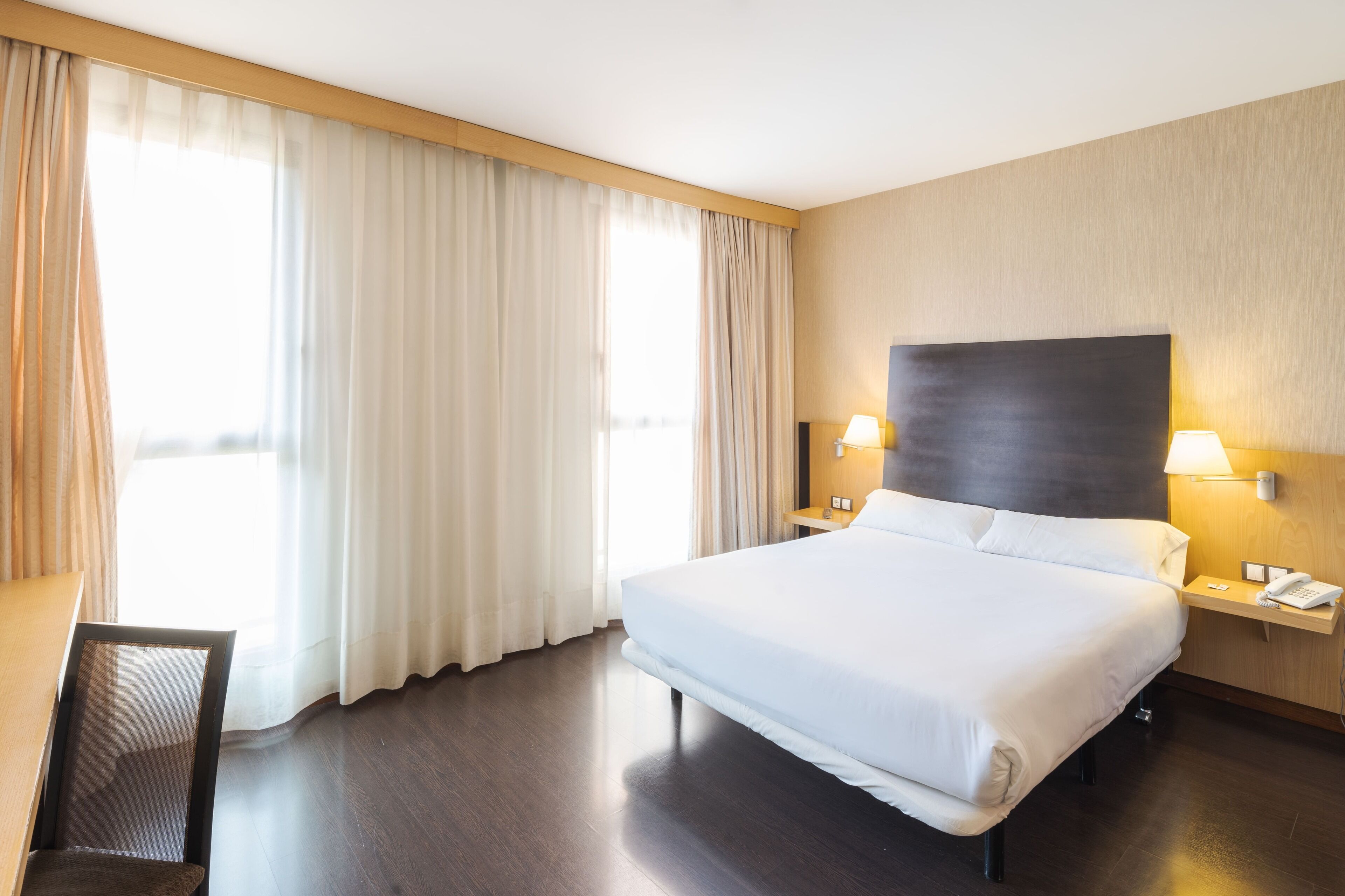 Habitación de hotel con una cama grande, dos mesitas de noche con lámparas, una silla y vistas al exterior a través de cortinas translúcidas.