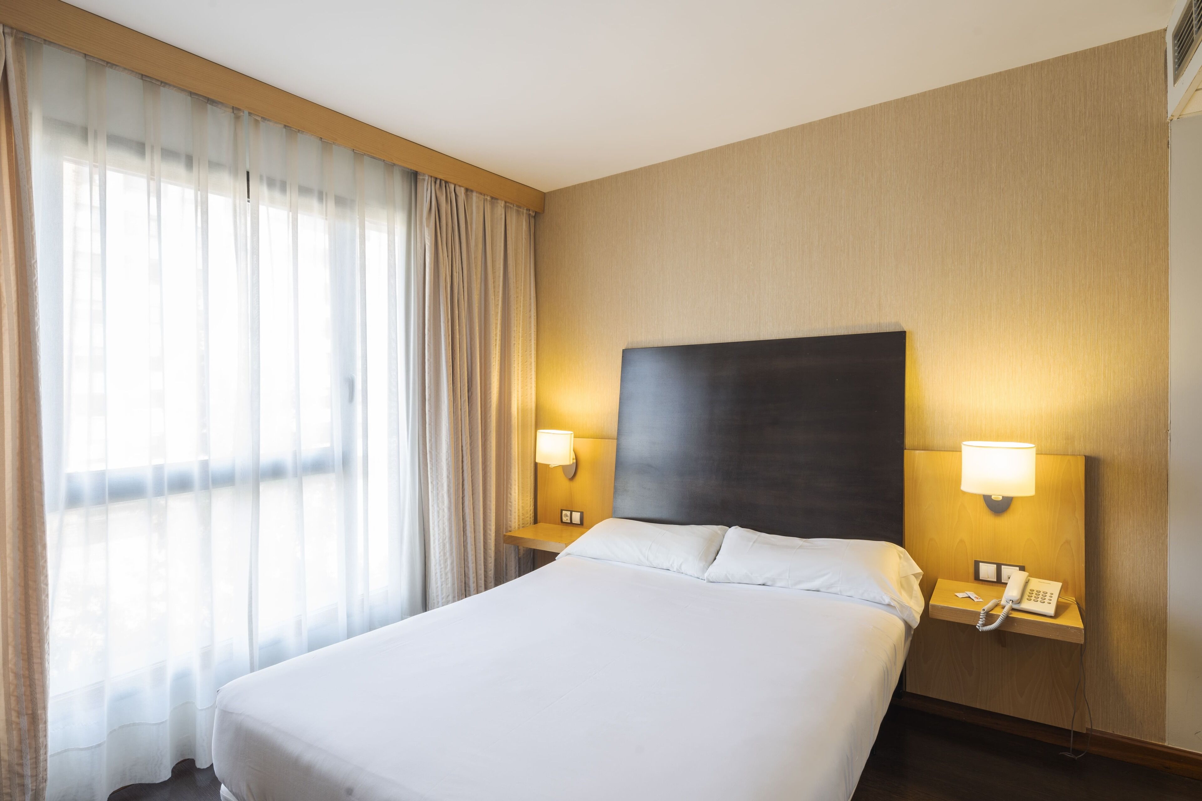 Una habitación de hotel con una cama grande, dos lámparas de noche, una mesita de noche con un teléfono y cortinas de gasa.
