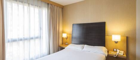 Una habitación de hotel con una cama grande, dos lámparas de noche, una mesita de noche con un teléfono y cortinas de gasa.