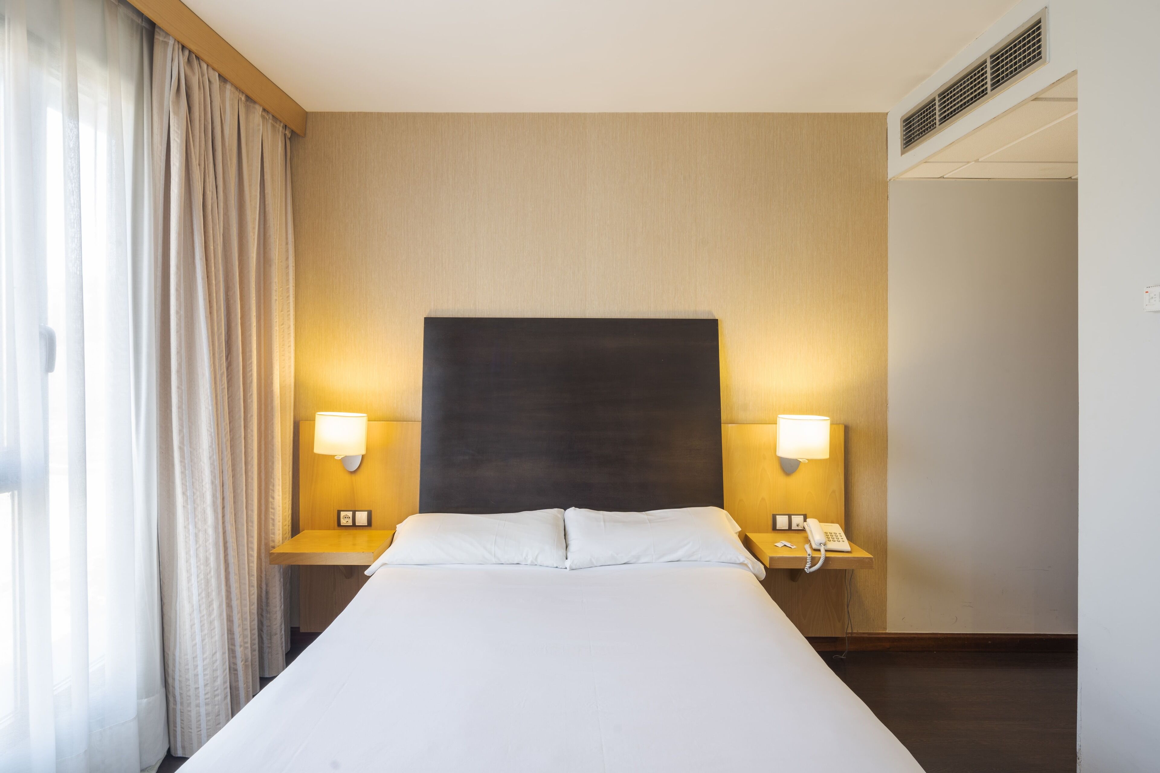 A hotel room with a large bed, two bedside tables with lamps, a telephone, and a window with curtains.