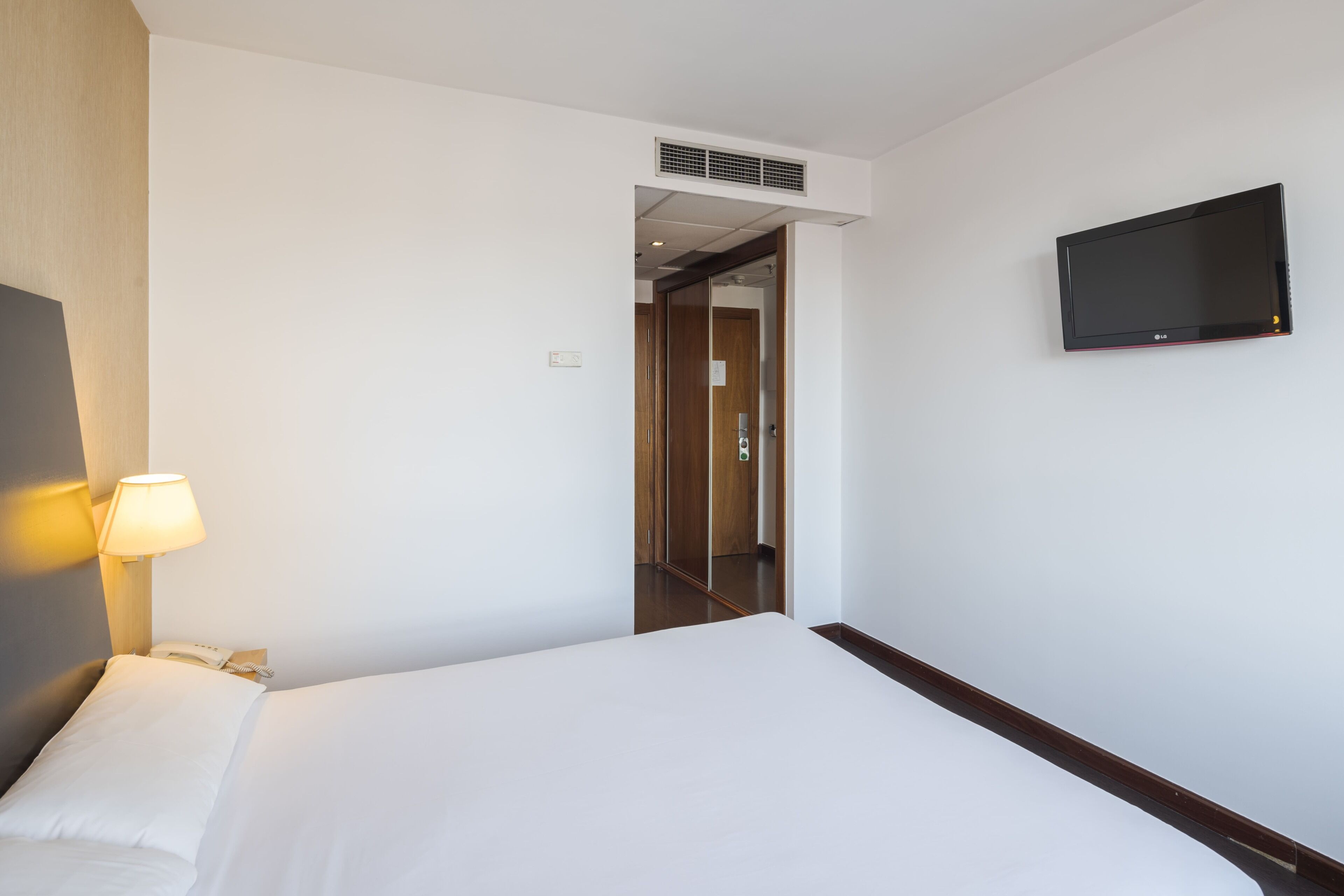 Habitación de hotel con cama, lámpara de noche, teléfono, televisor montado en la pared y puerta que da a otra habitación.