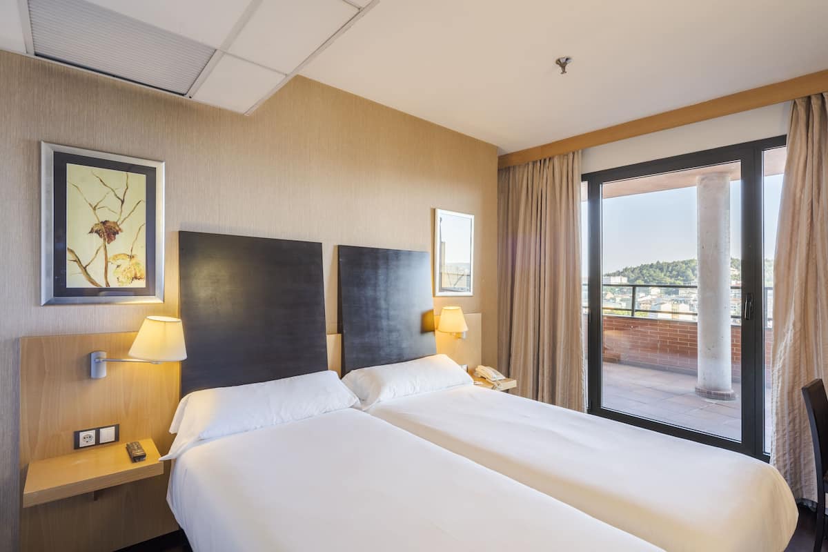 Une chambre d’hôtel avec deux lits, une table de chevet avec une lampe, un téléphone et une fenêtre donnant sur un immeuble et des arbres.