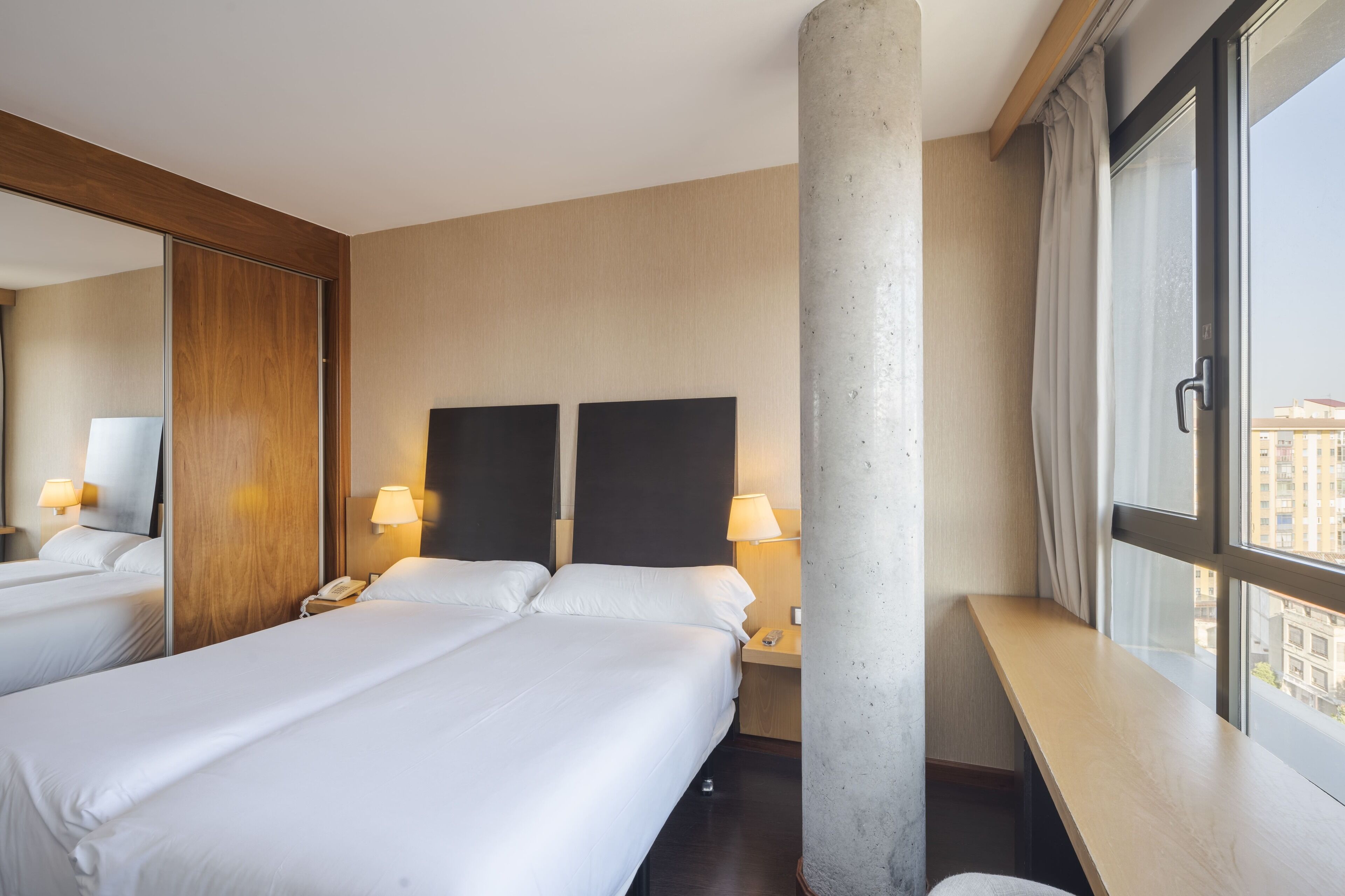 Une chambre d’hôtel avec deux lits, un grand miroir, un banc en bois et une fenêtre donnant sur la ville.